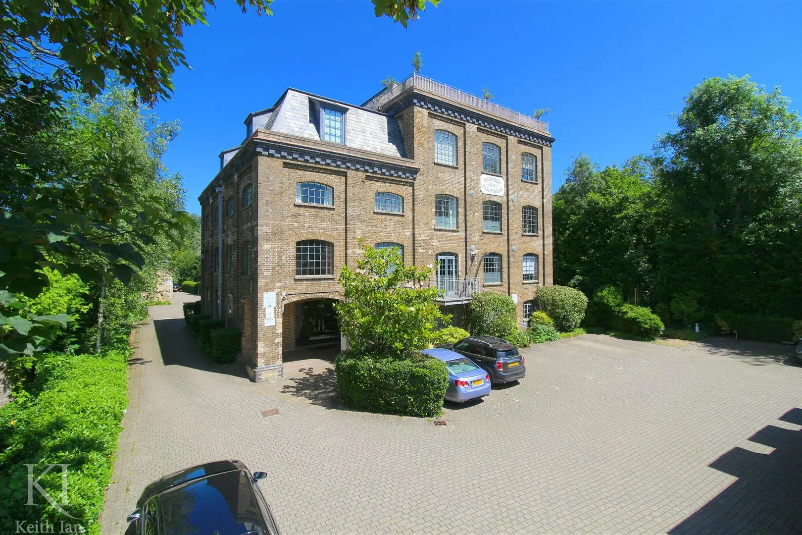 Period brick building with mansard roof and multiple sash windows, surrounded by mature trees and parked cars on a paved driveway