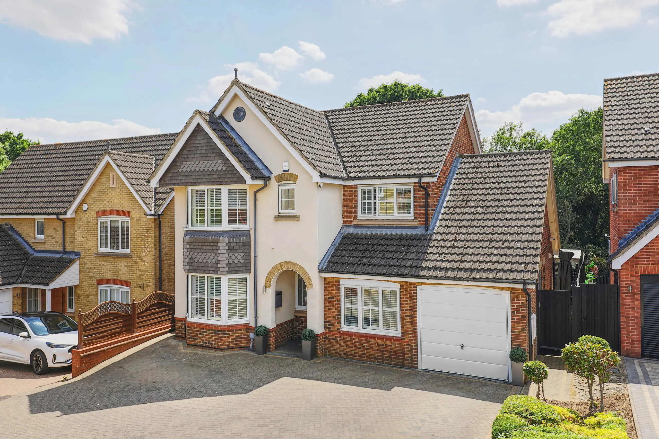 Modern two-storey detached house with cream and red brick exterior, white garage door, and dark tiled roof, set on a paved driveway with landscaped front garden