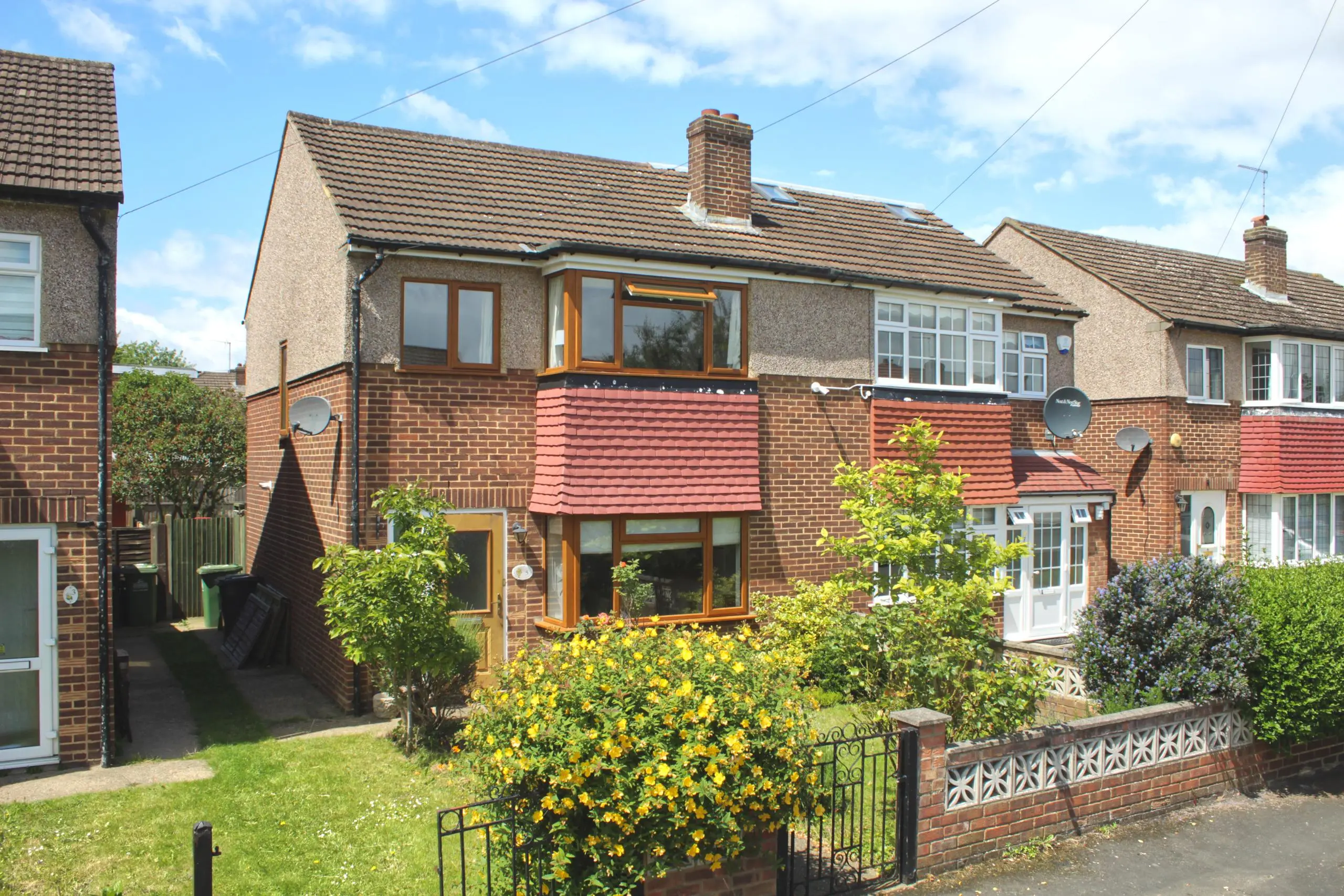 Semi-detached brick house with rendered upper storey, red tiled roof, brown-framed windows, and front garden with flowering shrubs and metal fence