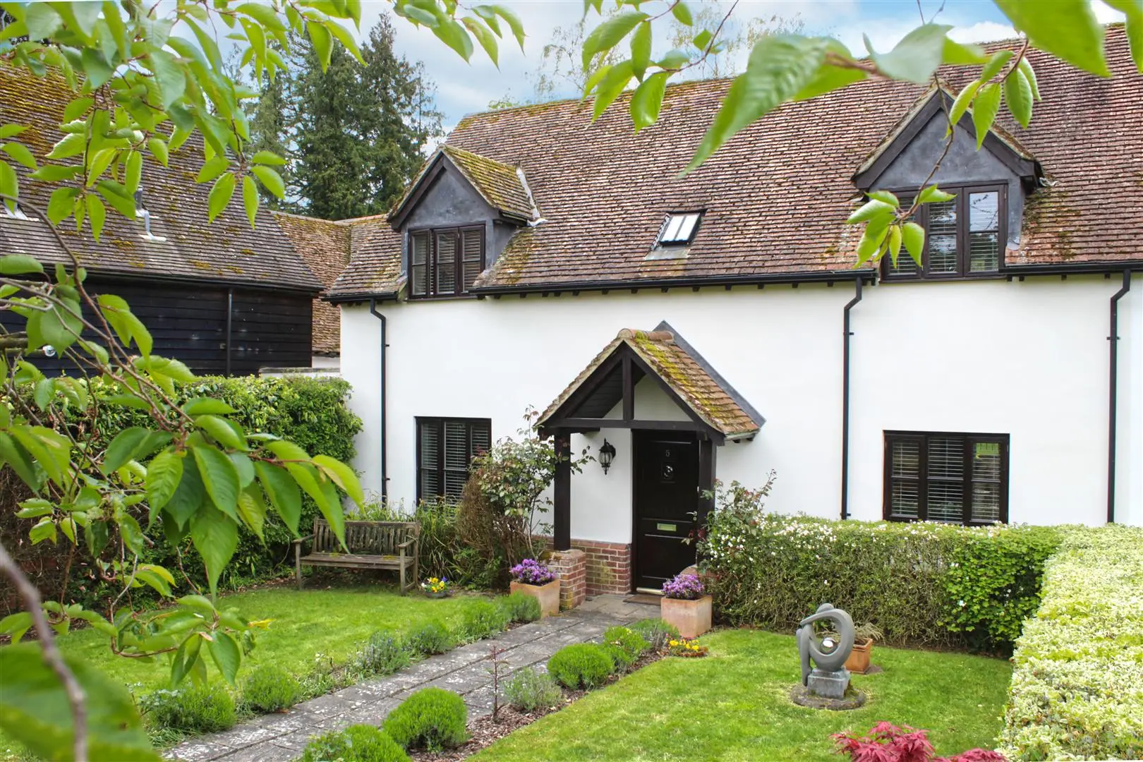 White rendered cottage with tiled roof, black door and shutters, brick chimneys, and manicured garden with lawn, hedges and planted borders