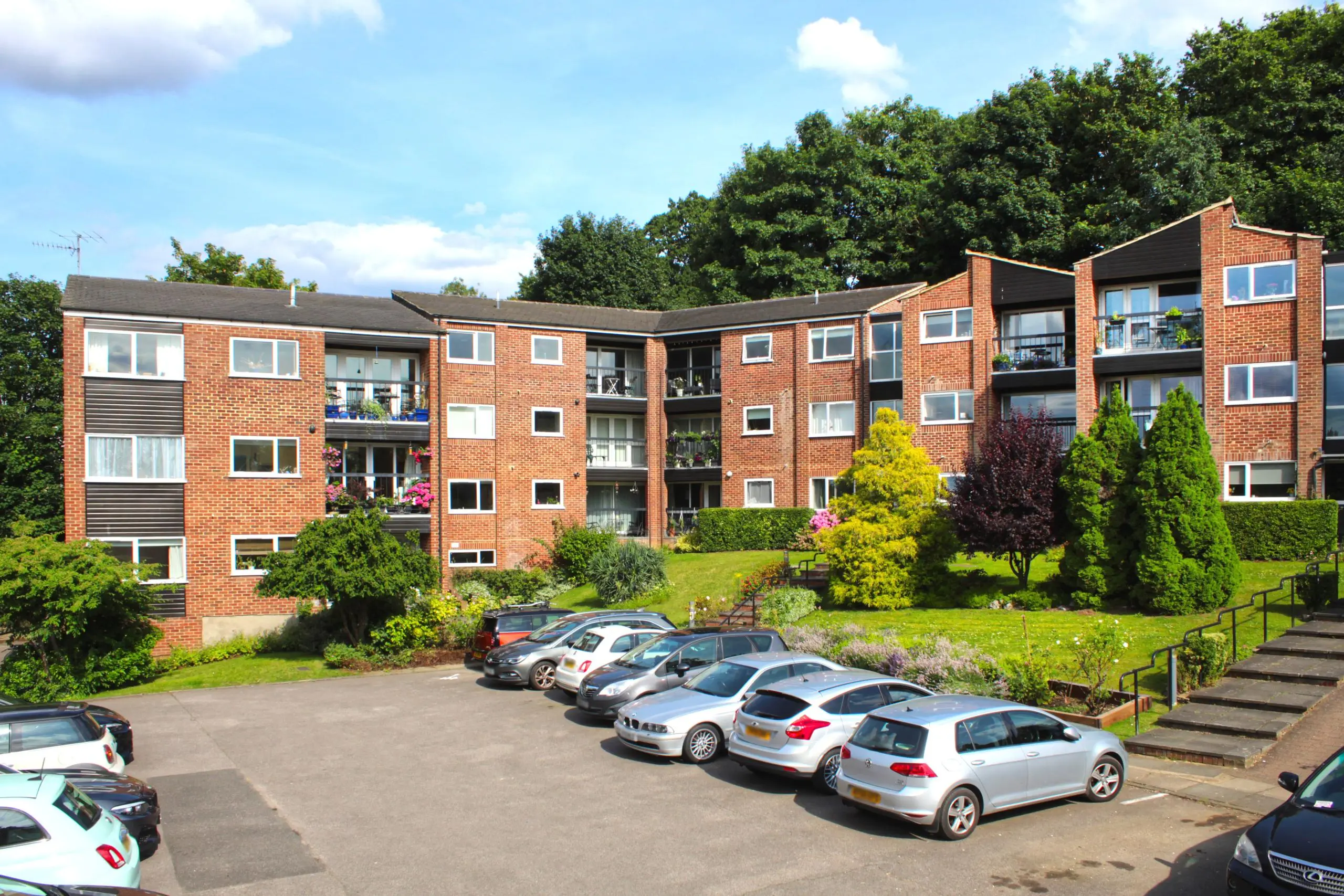 Red brick apartment building with multiple storeys, balconies, and surrounding landscaped gardens with shrubs and trees. Parked cars visible in foreground.