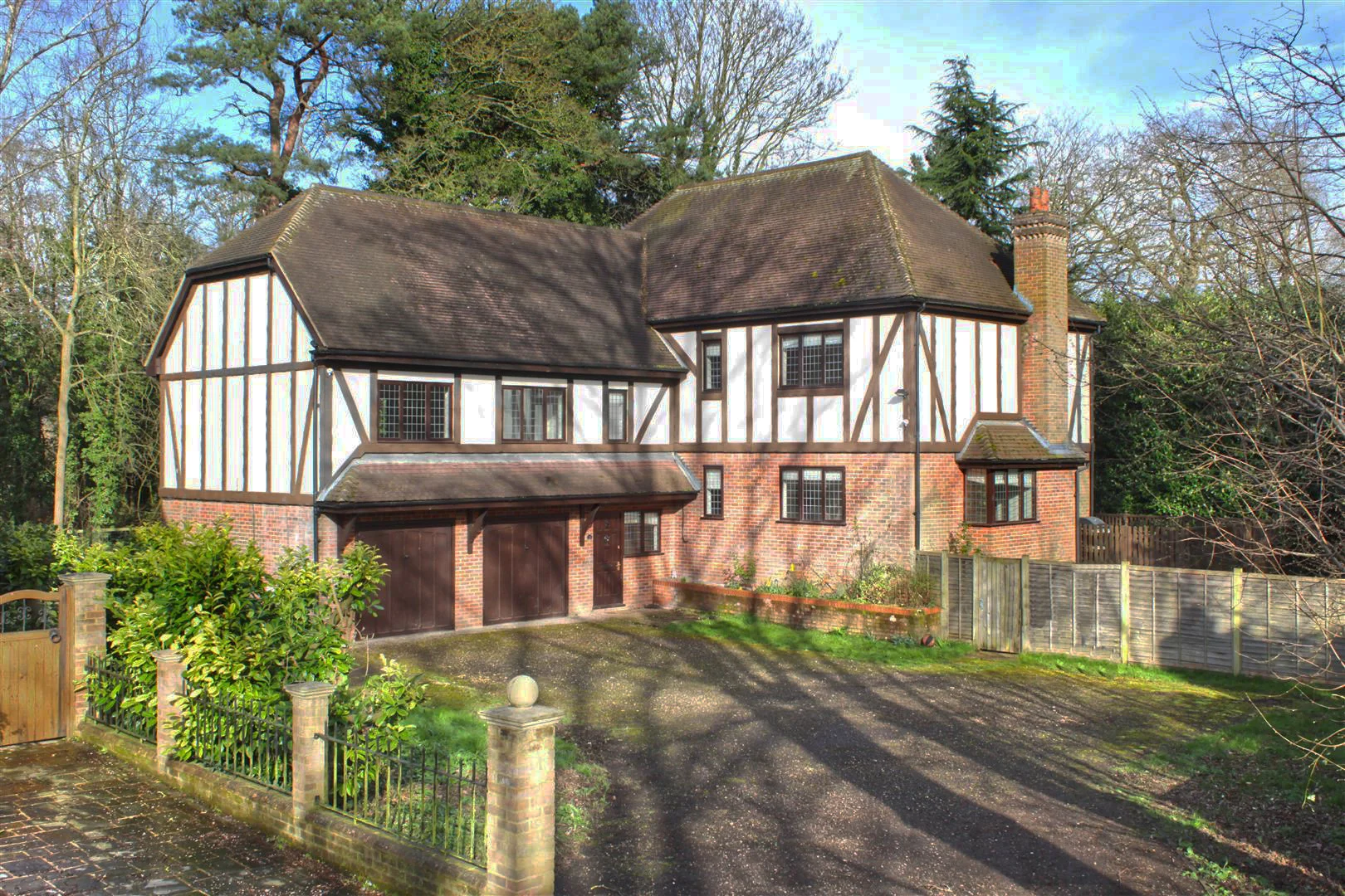 Tudor-style house with white-painted timber framing and brick lower storey, dark tiled roof, brick chimney, double garage, driveway and surrounding trees