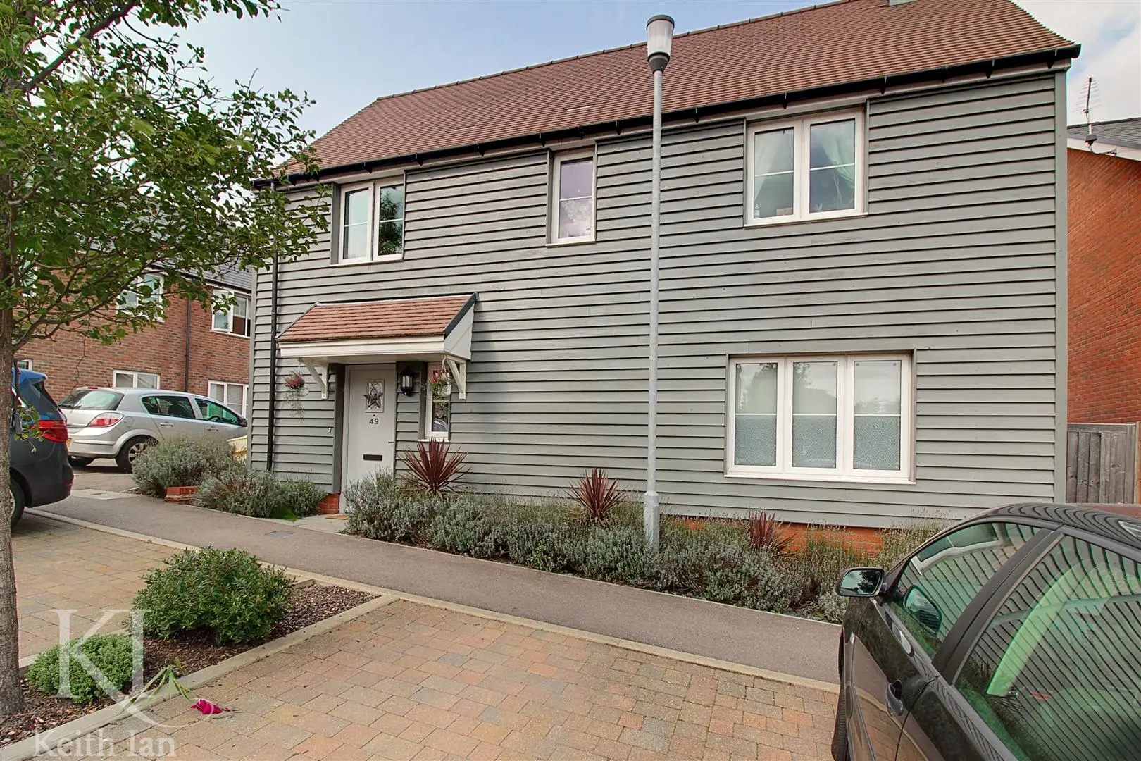 Modern house with grey horizontal cladding, brown tiled roof, white-framed windows, and number 49 on the front door. Parked cars visible on the street.