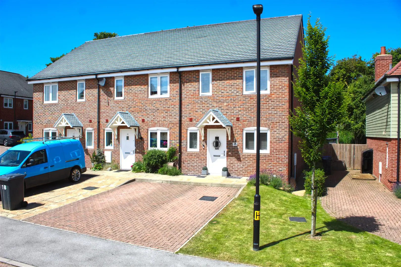 Brick terraced houses with white doors and pitched slate roof, brick paved driveway, parked blue van, clear sky