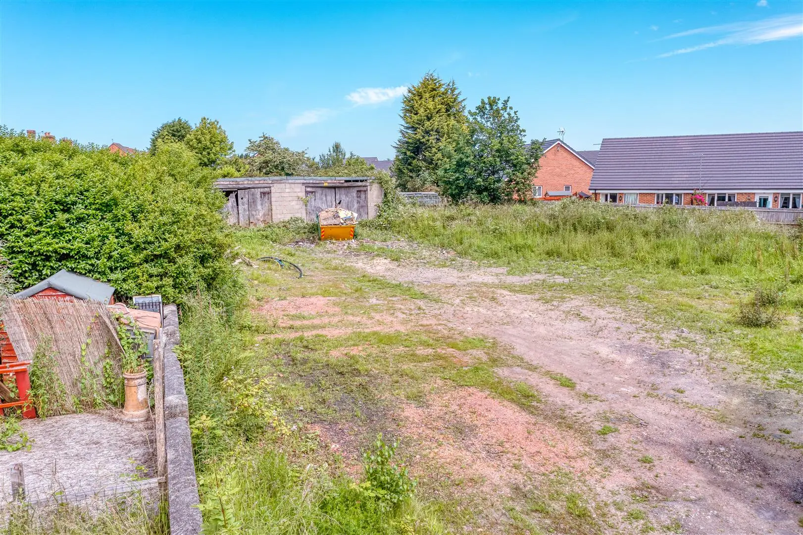 Large overgrown garden with wooden sheds, orange skip, and brick houses visible in background