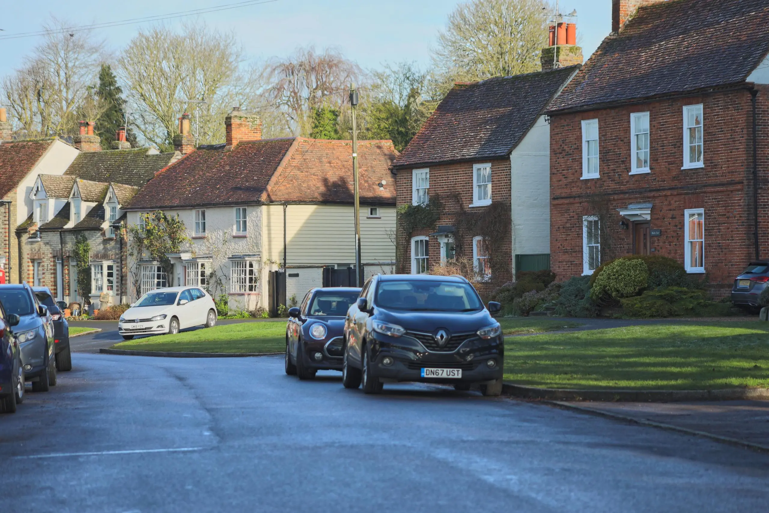Row of brick and rendered cottages with pitched roofs, parked cars on road, bare trees and clear sky