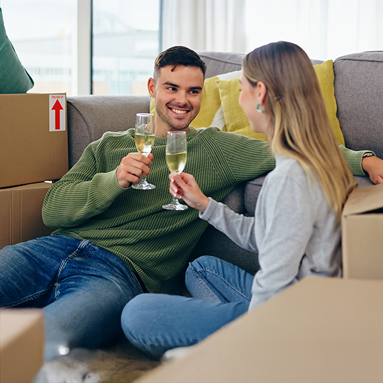 A couple sit on a sofa clinking glasses, surrounded by moving boxes