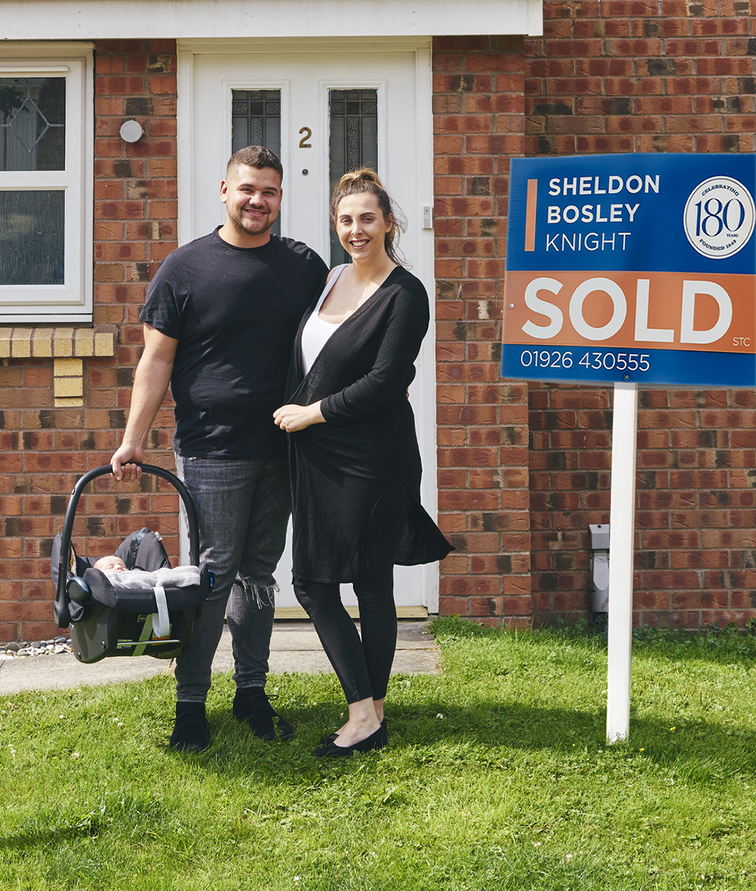 Two homeowners stand outside a new house with a Sheldon Bosley Knight sold board on the grass