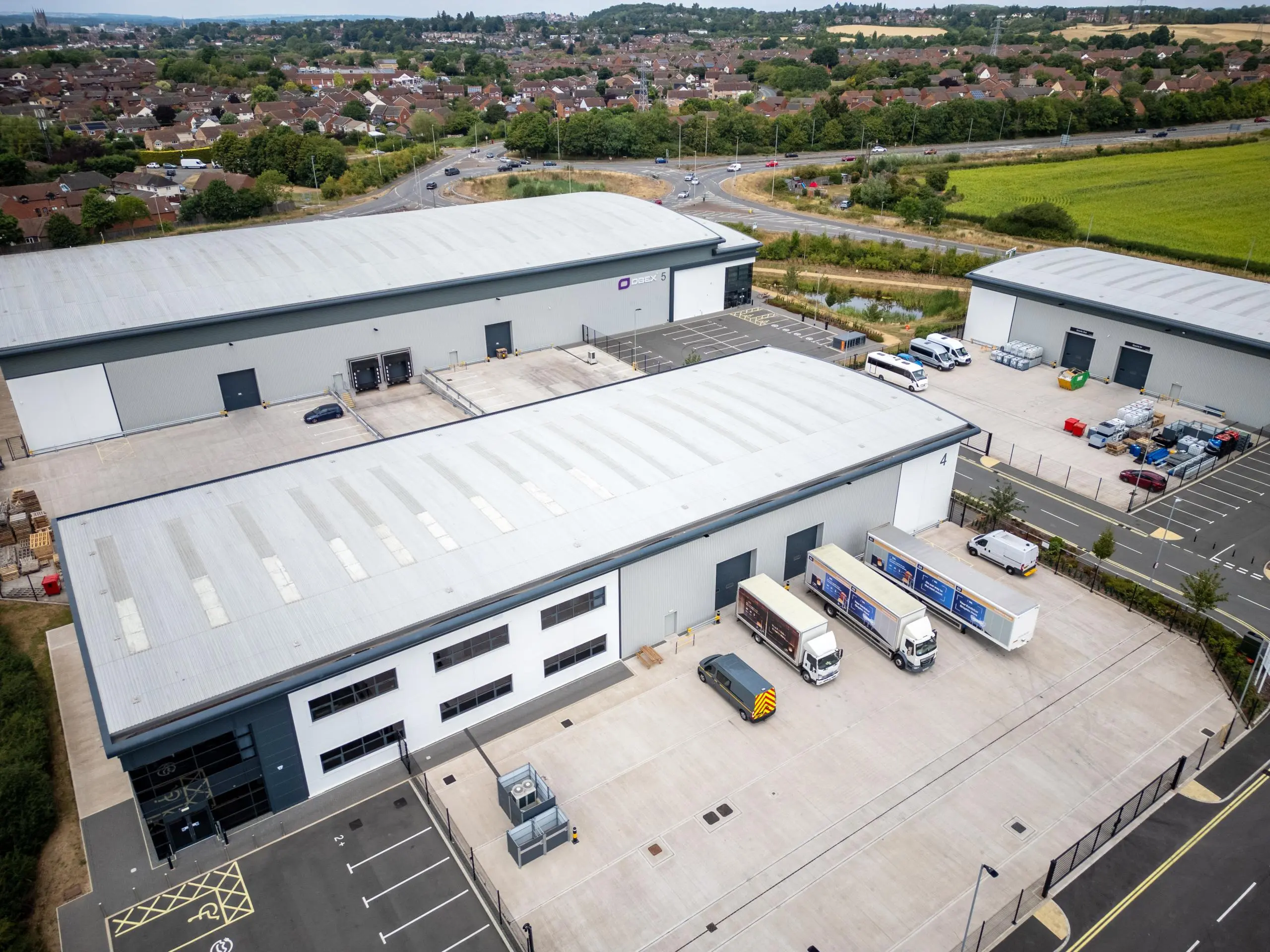 Aerial view of modern industrial warehouse buildings with white metal roofs, parked vehicles and loading areas, surrounded by residential suburbs and green fields