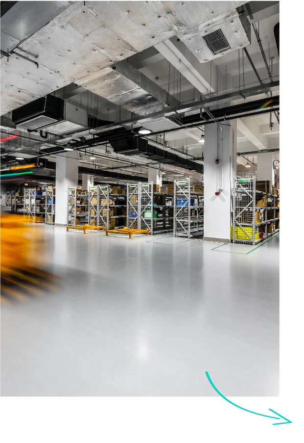 Industrial warehouse interior with exposed concrete ceiling, metal ducting, white support columns, and metal storage cages with yellow safety barriers
