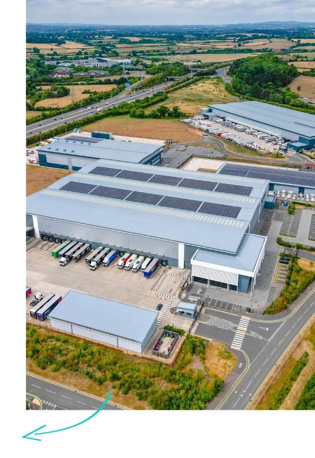 Aerial view of industrial warehouse complex with solar panels on roof, loading bays with vehicles, and surrounding agricultural fields and motorway