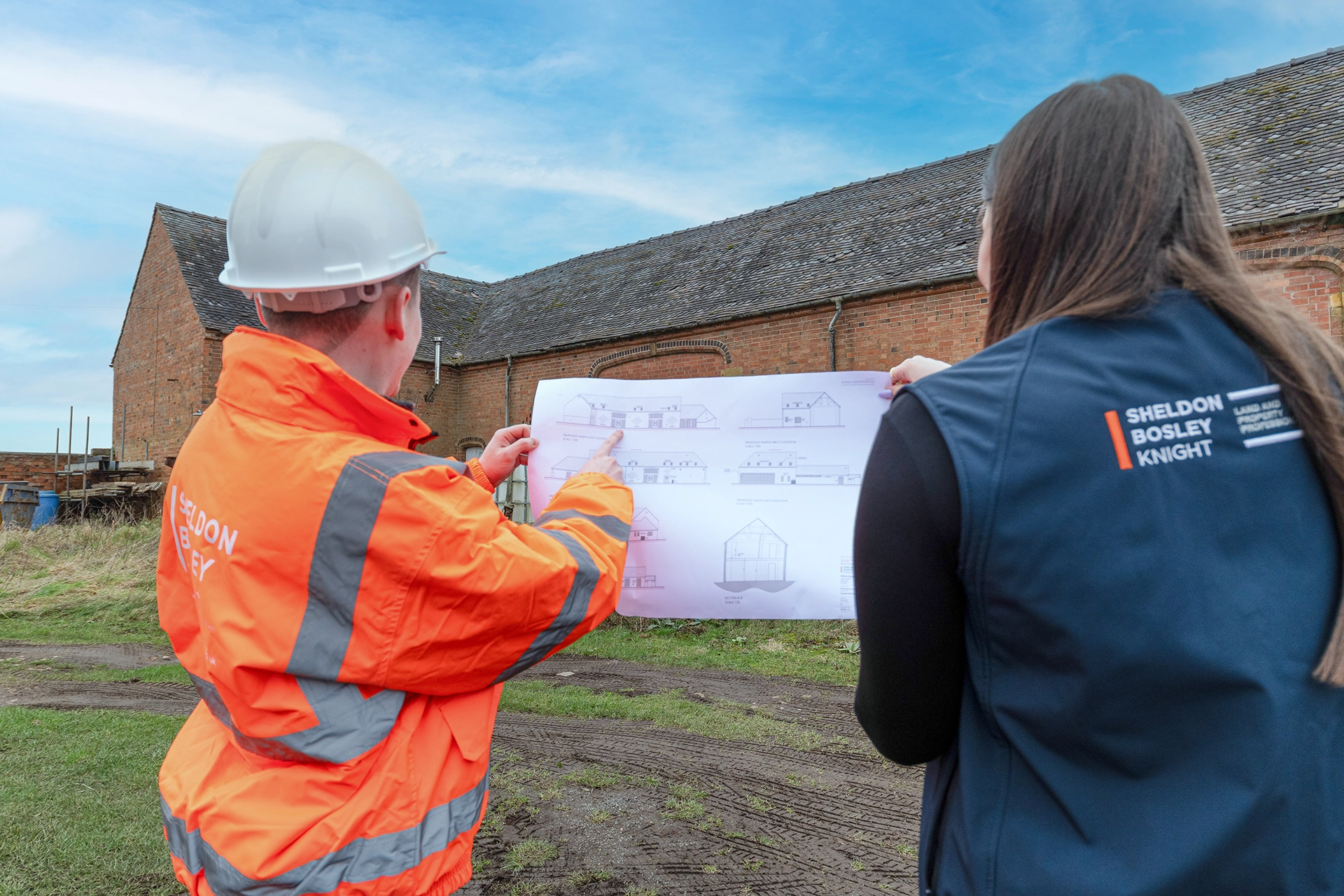 Two people viewing architectural plans outdoors in front of a brick building. Person on left wears white hard hat and orange high-visibility jacket with Sheldon Bosley Knight branding. Person on right wears navy jacket with Sheldon Bosley Knight logo. Building sketches visible on the plans.
