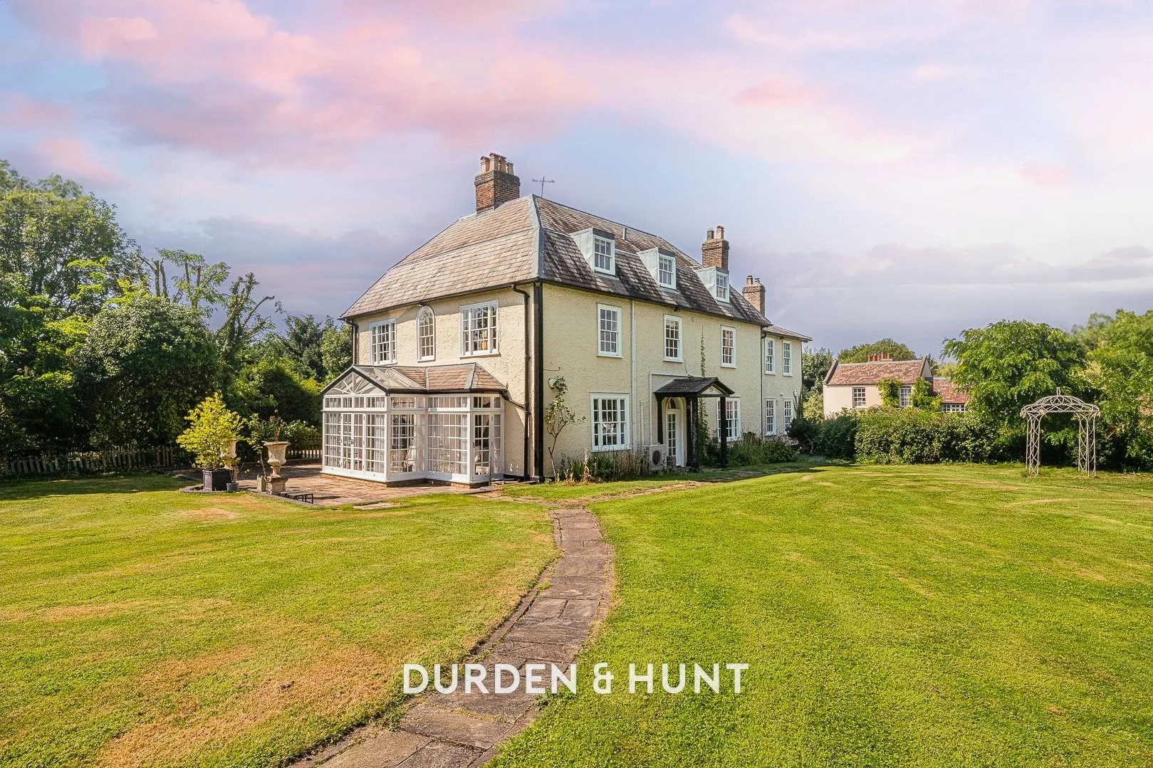Cream-coloured country house with slate roof, brick chimneys, and glazed extension, set in landscaped grounds with lawn, trees, and metal gazebo. Durden & Hunt branding visible.