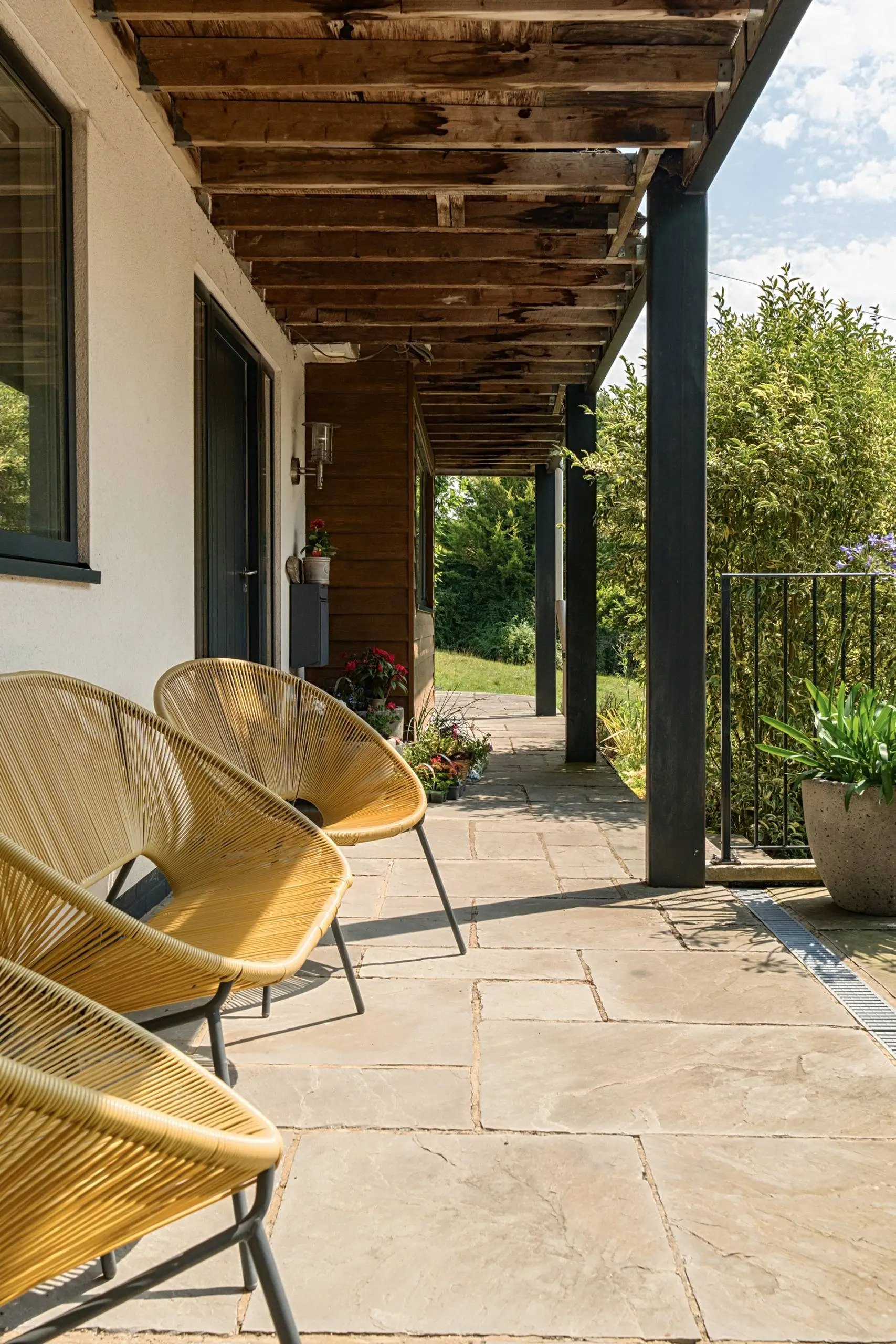 Covered patio with wooden pergola, two yellow woven chairs, stone paving, potted plants, and views over green garden and countryside
