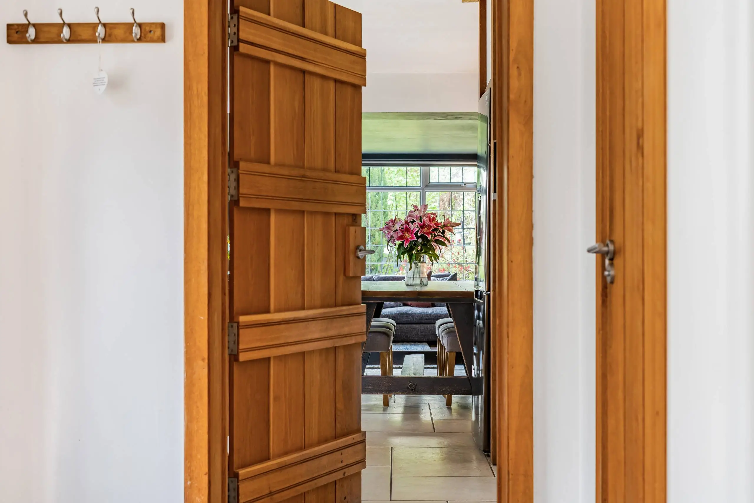 View through open wooden doors into a room with wooden flooring, a window with green blind, pink flowers in a vase on the windowsill, and furniture including chairs and a dark table