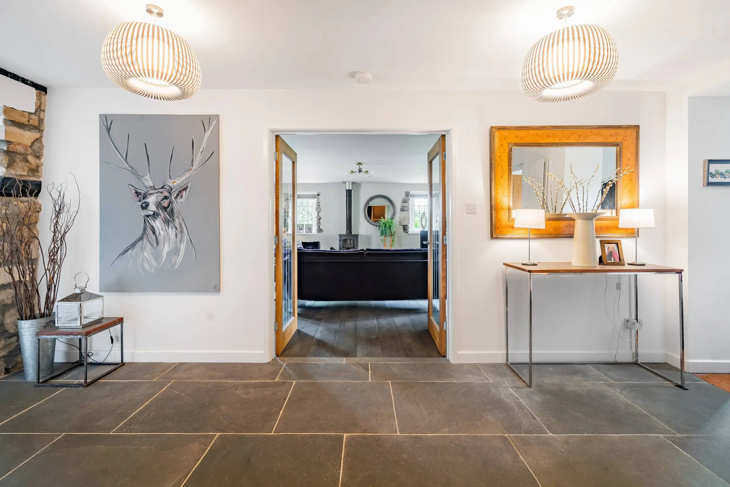 Modern hallway with slate flooring, two pendant lights, deer artwork on grey canvas, wooden console table with decorative items, gold-framed mirror, and open wooden doors leading to living room with black fireplace