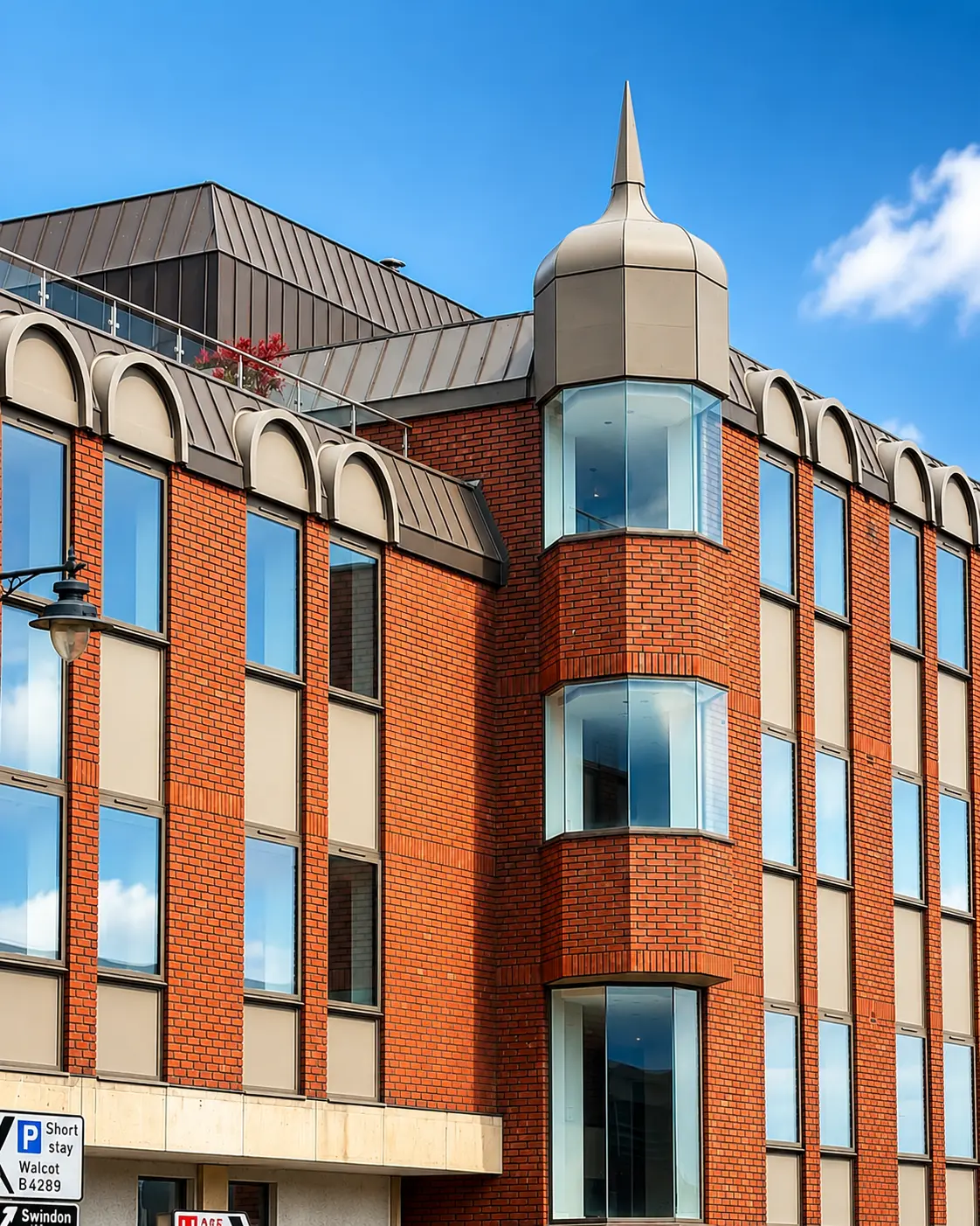 Red brick building with arched windows, metal roof, and distinctive domed turret with spire. Street signs visible at base including Walcot and Swindon.