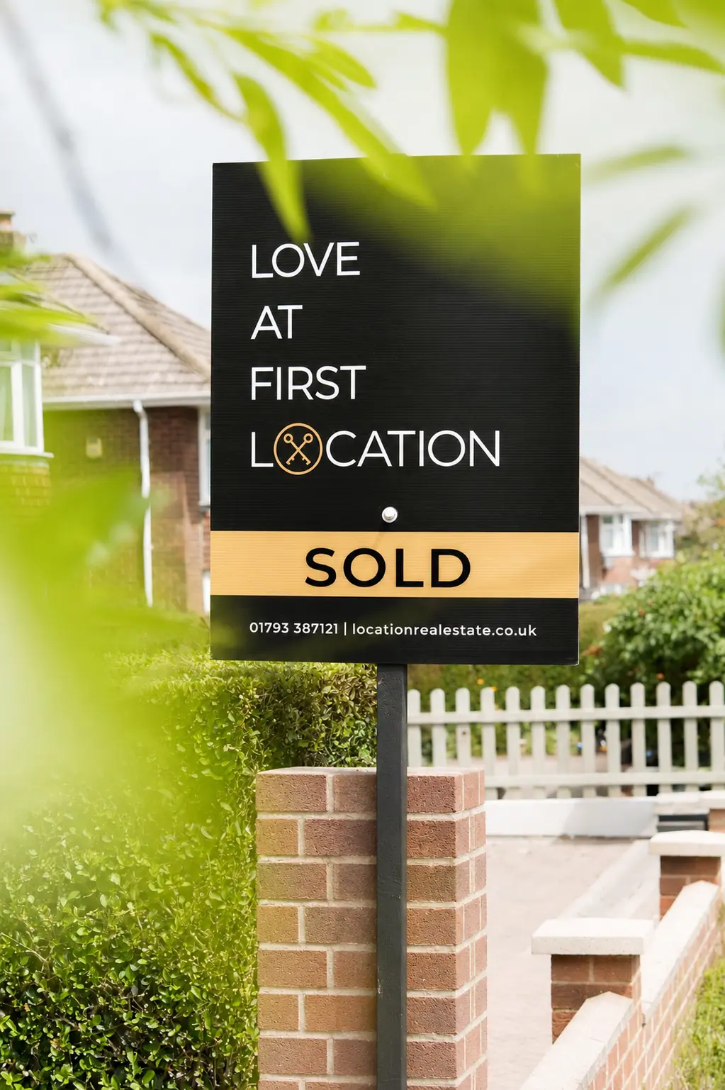Estate agent sold sign reading 'Love at first location' with 'SOLD' banner, phone number 01793 387121 and website locationrealestate.co.uk, mounted on brick pillar in front garden