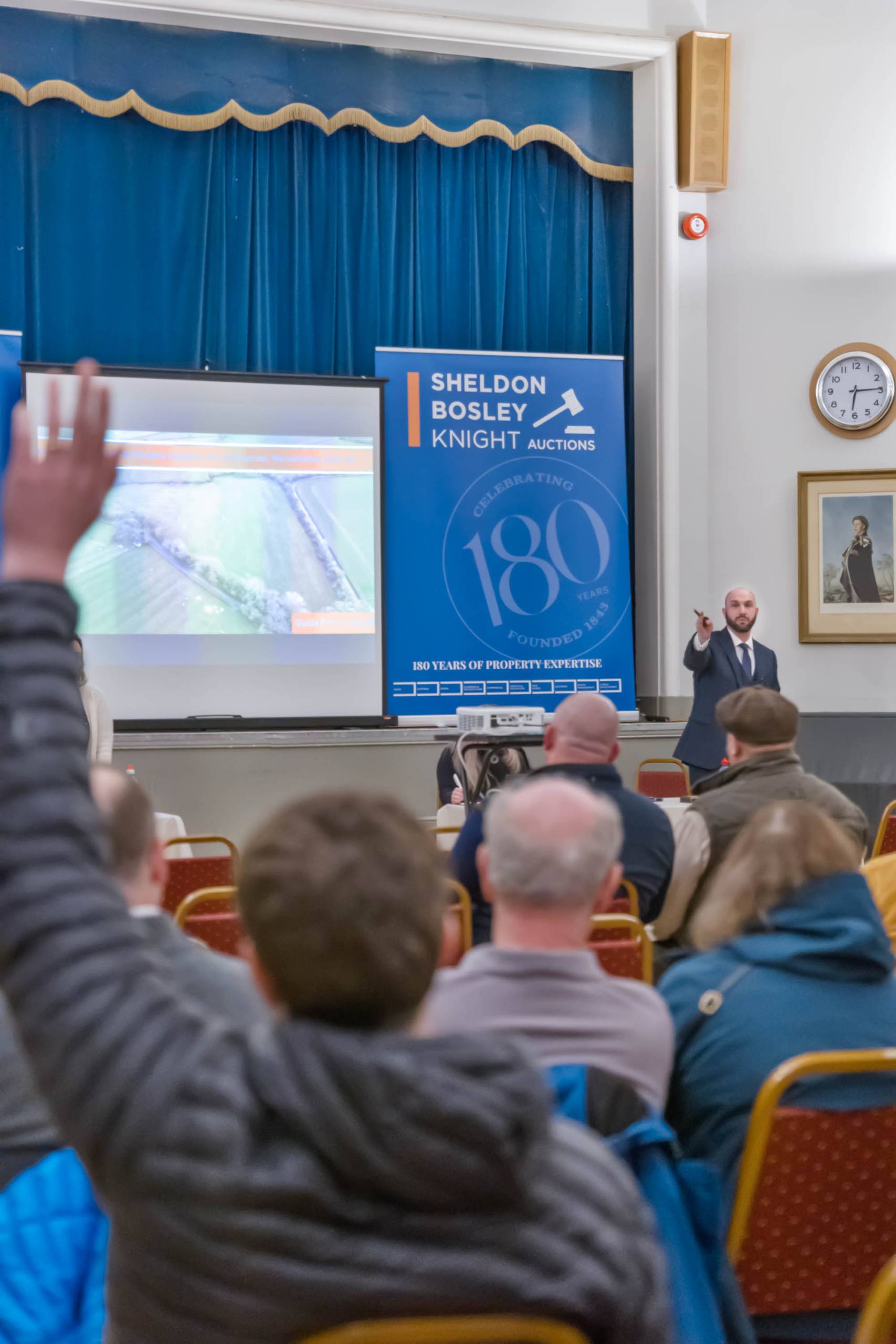 Buyers raise hands in a Sheldon Bosley Knight auction room. An auctioneer stands at the front of the room