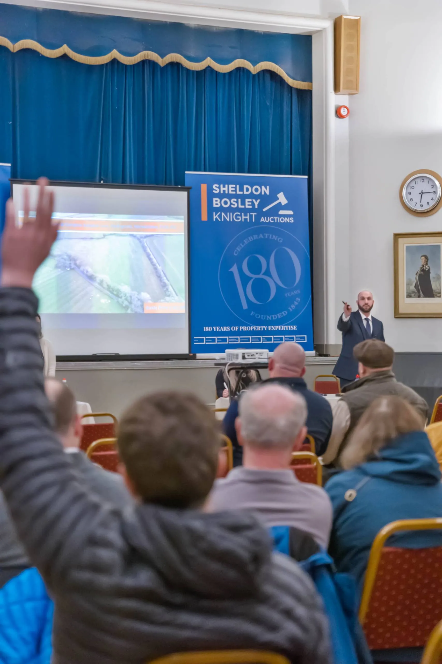 Buyers raise hands in a Sheldon Bosley Knight auction room. An auctioneer stands at the front of the room