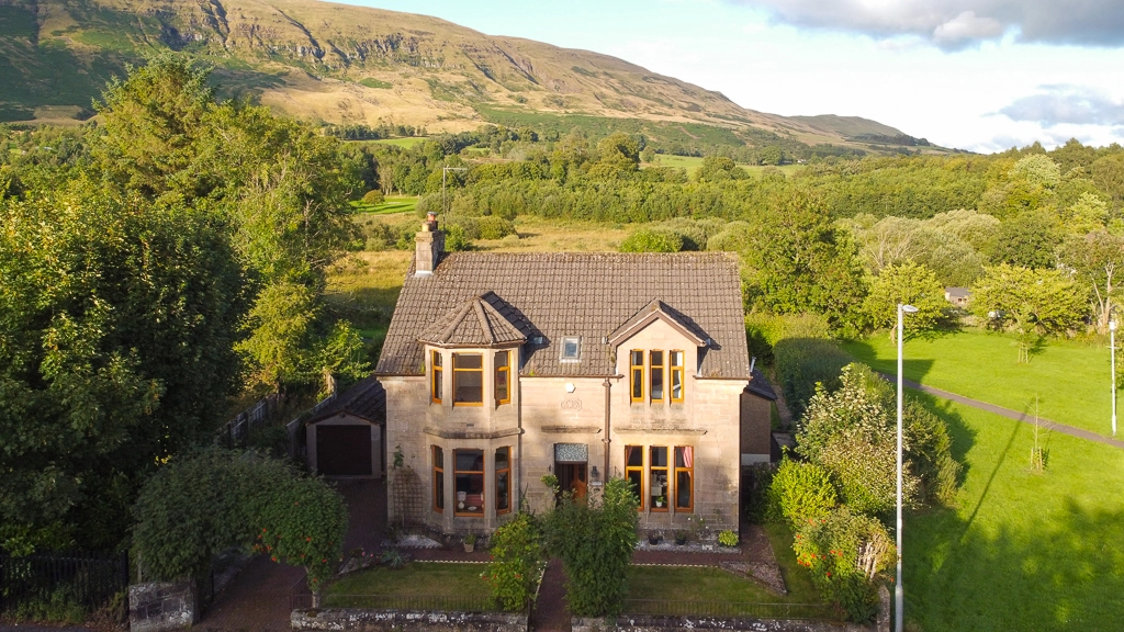 Stone house with brown tiled roof and yellow-framed windows, surrounded by trees and green fields, with mountains in the background