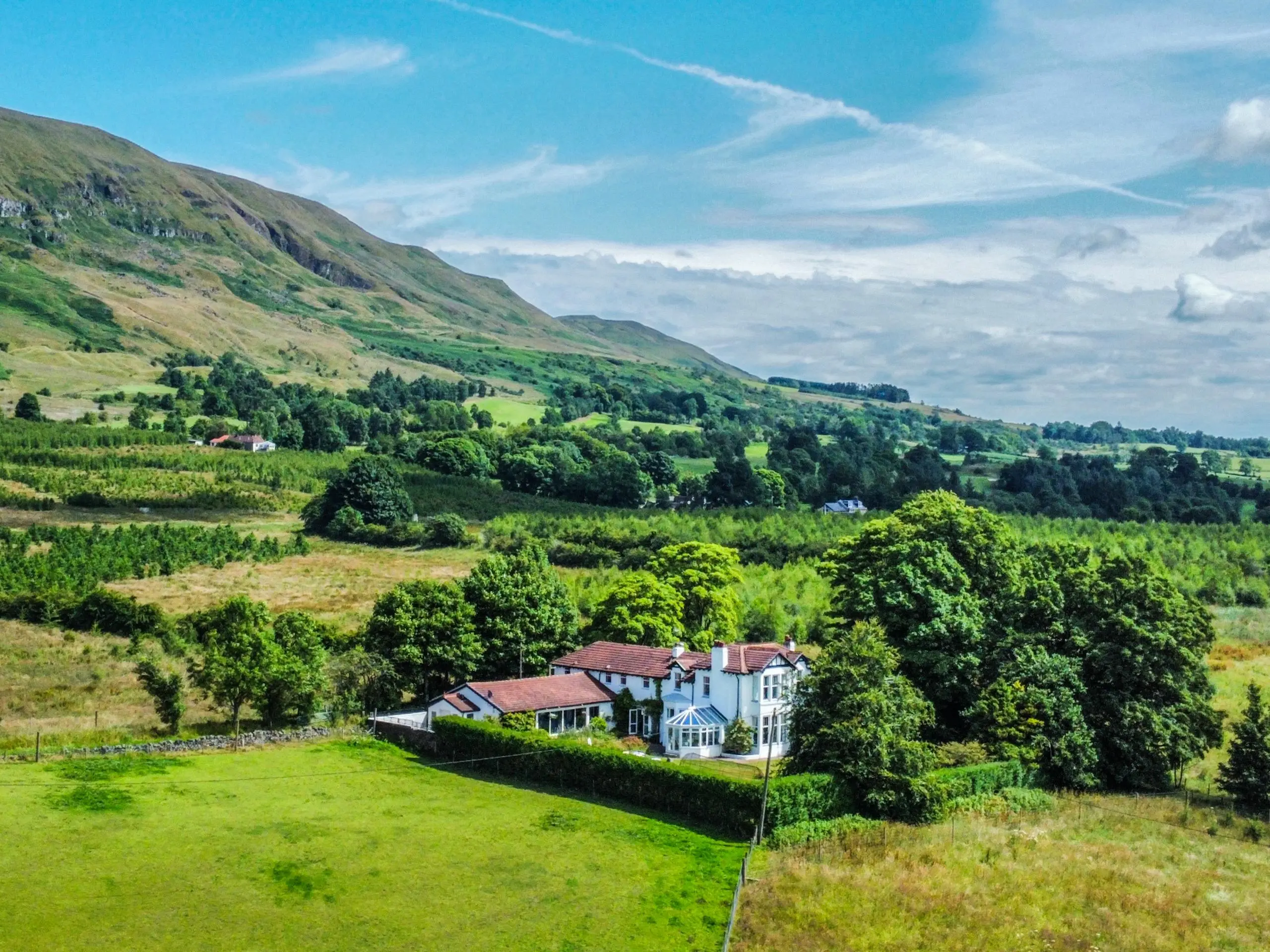 Country house with white and stone exterior surrounded by hedges and trees, set in rolling green valley with mountains in background