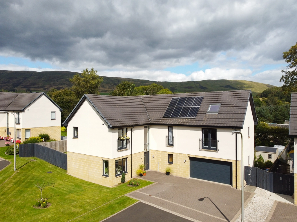 Modern two-storey house with white render and stone base, dark roof with solar panels, integrated garage, driveway, and landscaped garden with rolling hills in background