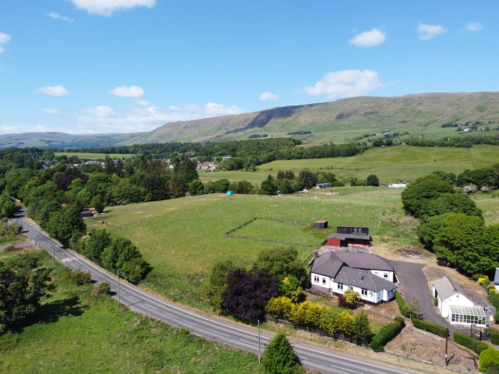 Aerial view of a white and grey house with outbuildings in a rural valley landscape, surrounded by green fields and hills