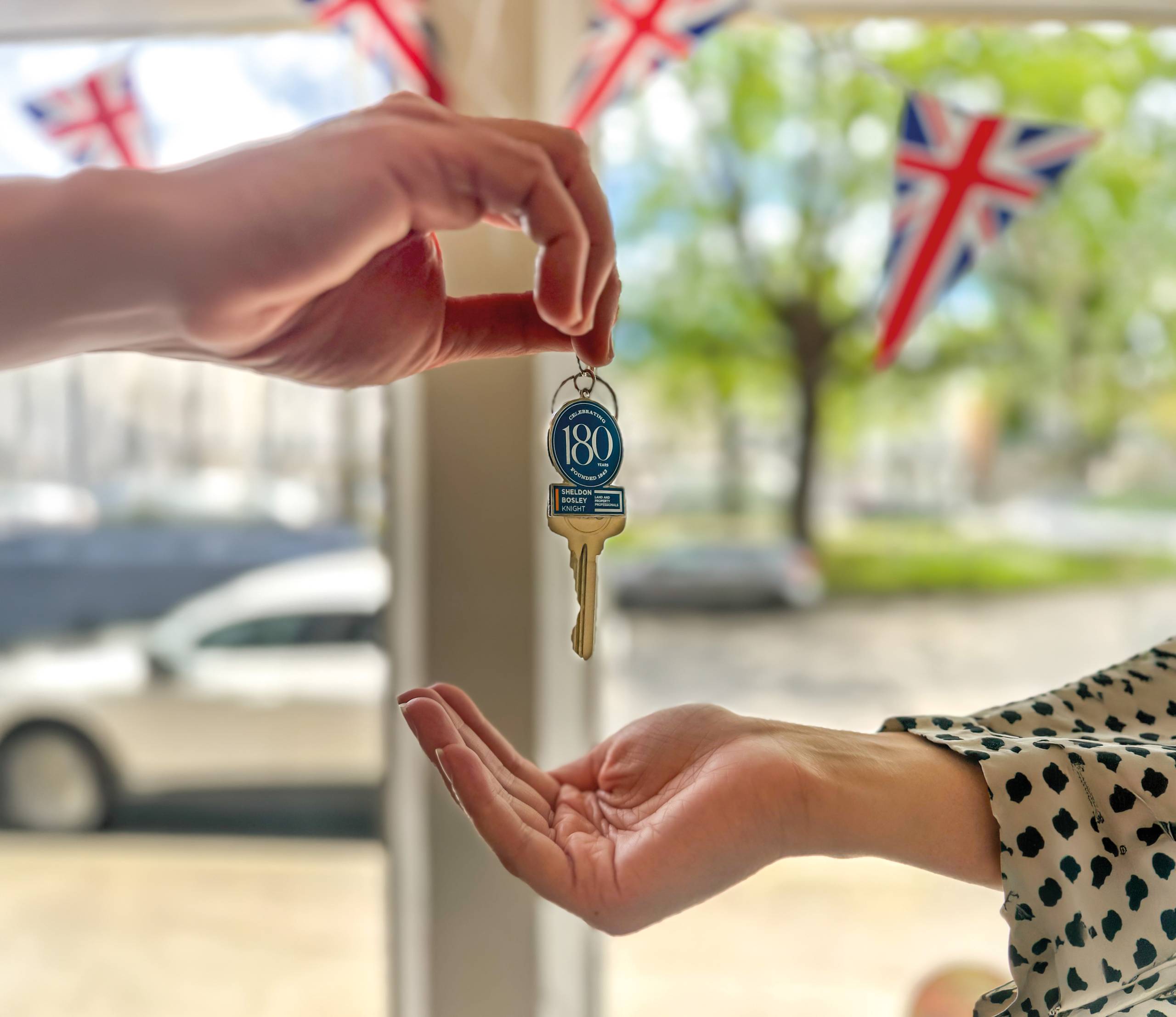 An estate agent's hands passing new keys to a buyer. The keys have Sheldon Bosley Knight branding