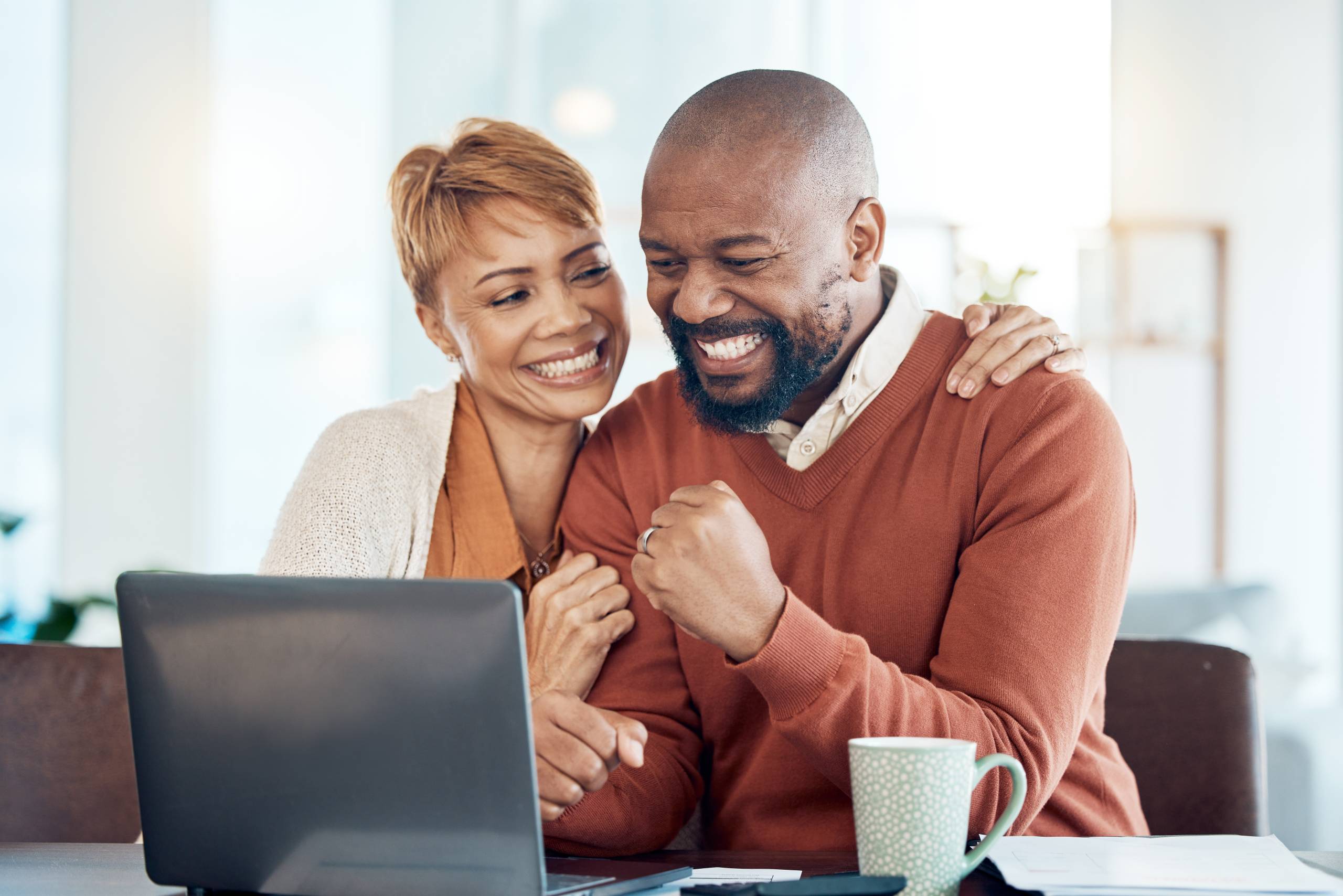 A couple sit behind a laptop screen, hugging and celebrating