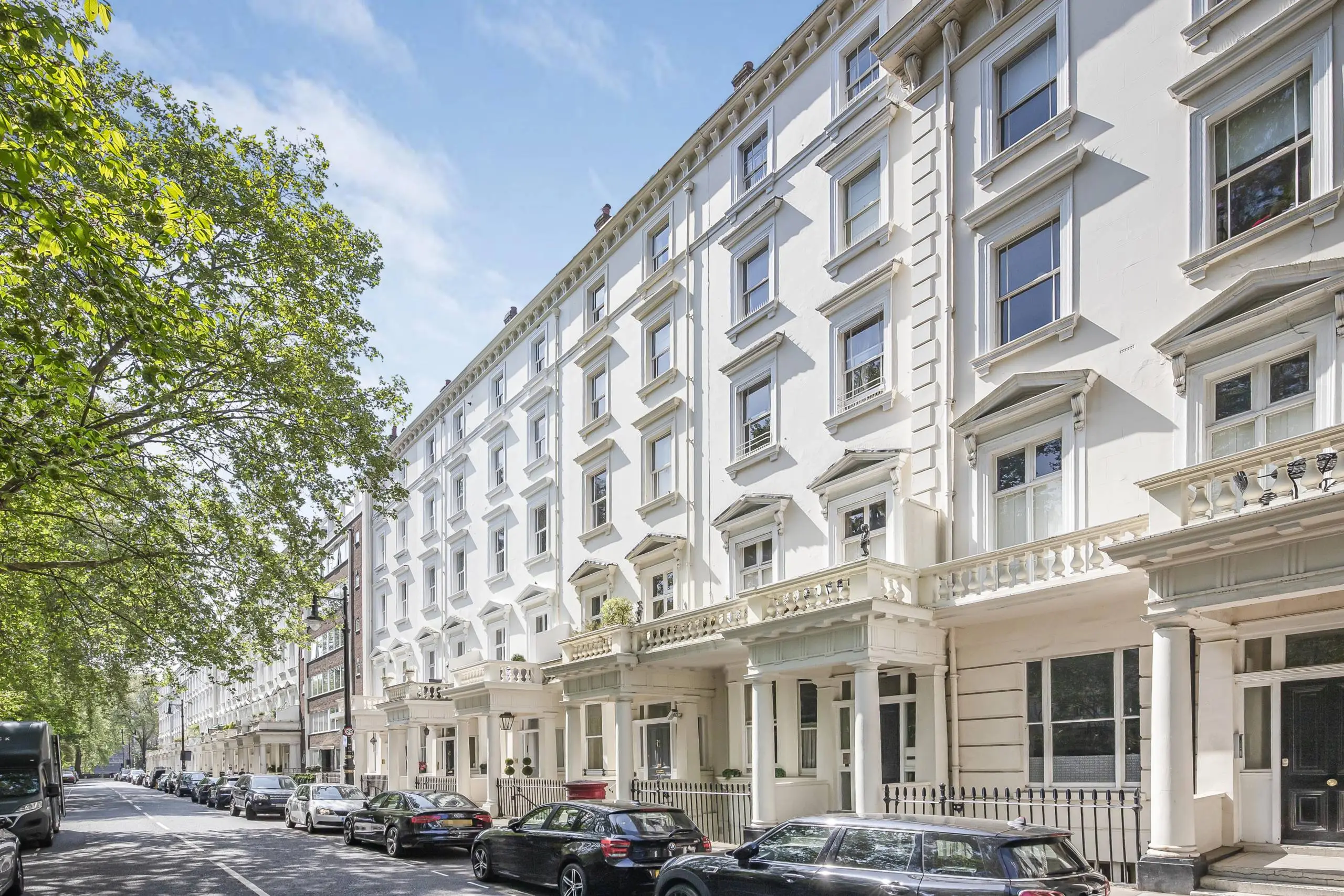 White stucco mansion block with multiple storeys, sash windows, and ground-floor colonnade, parked cars on street, tree foliage on left
