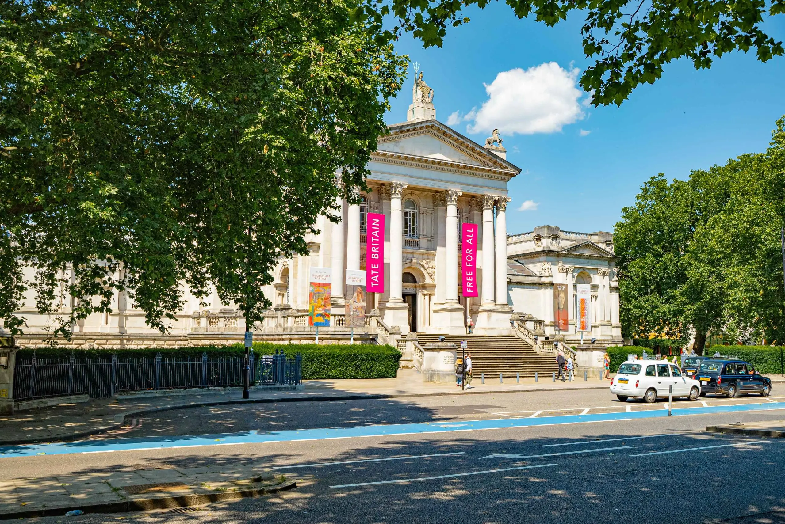 Neoclassical building with white columns and dome, flanked by pink banners reading "FREE BRITAIN" and "FREE FOR ALL", surrounded by trees and parked cars