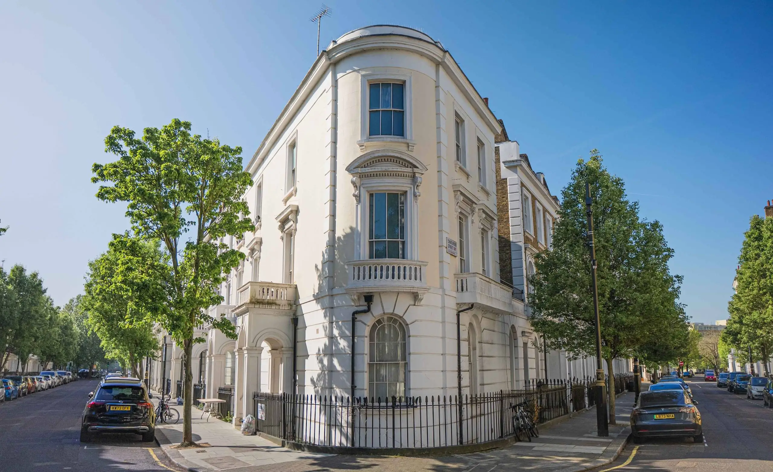Cream-coloured corner townhouse with curved bay windows, ornamental balustrade, and black iron railings, set on a tree-lined street with parked cars
