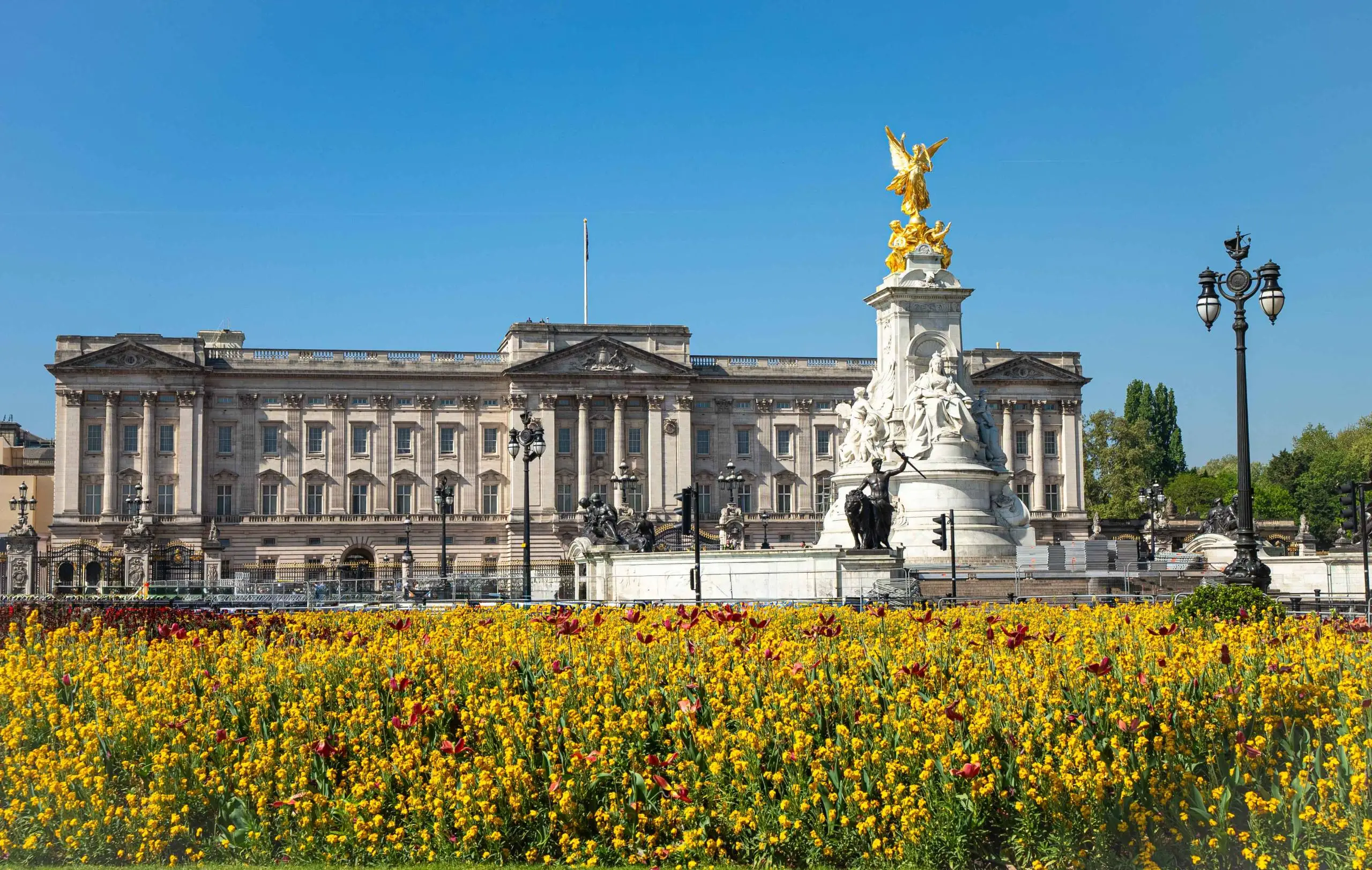 Buckingham Palace with a golden winged statue monument in the foreground, surrounded by yellow and red flowers