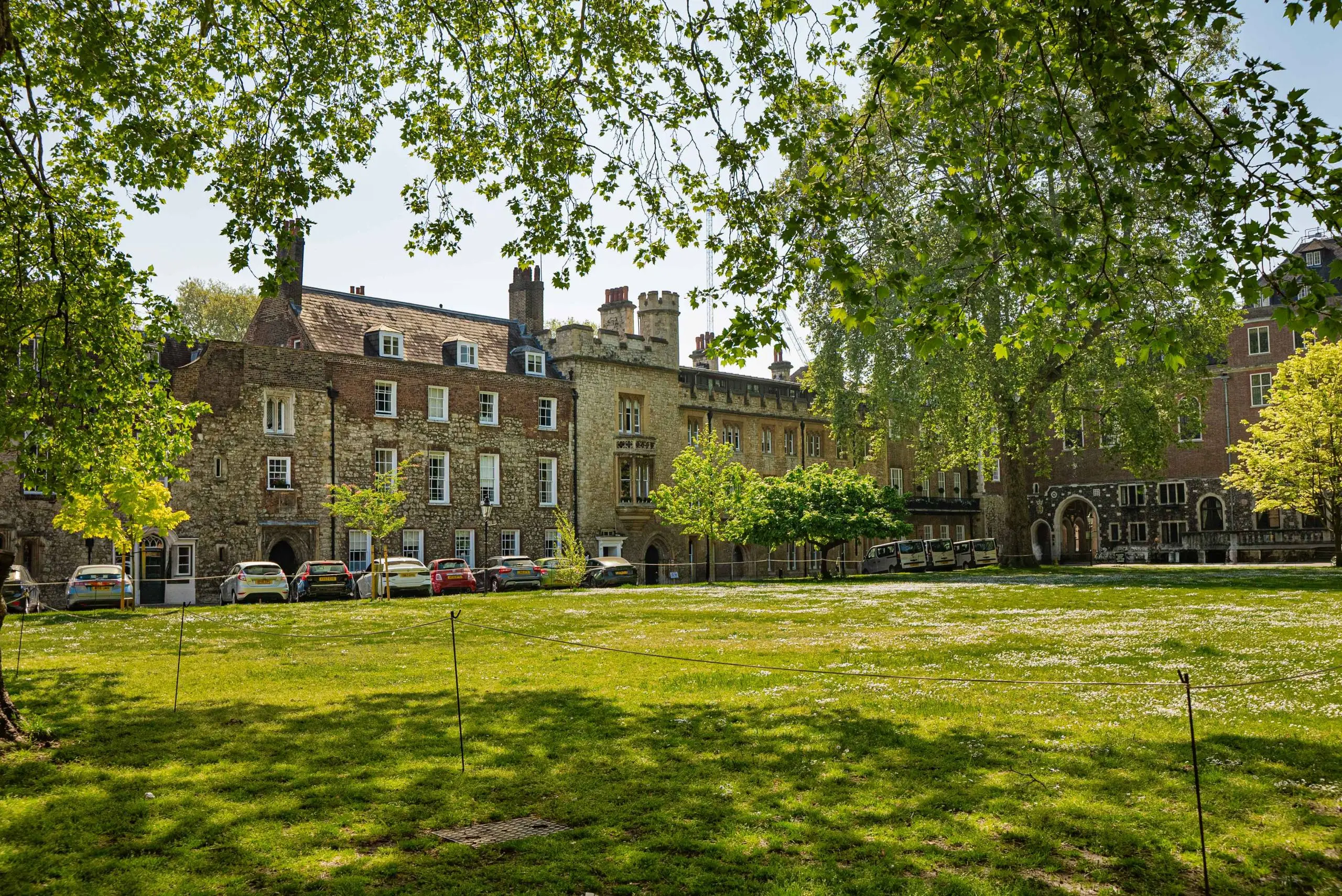 Historic stone buildings with crenellated towers surrounding a green courtyard with parked cars and mature trees