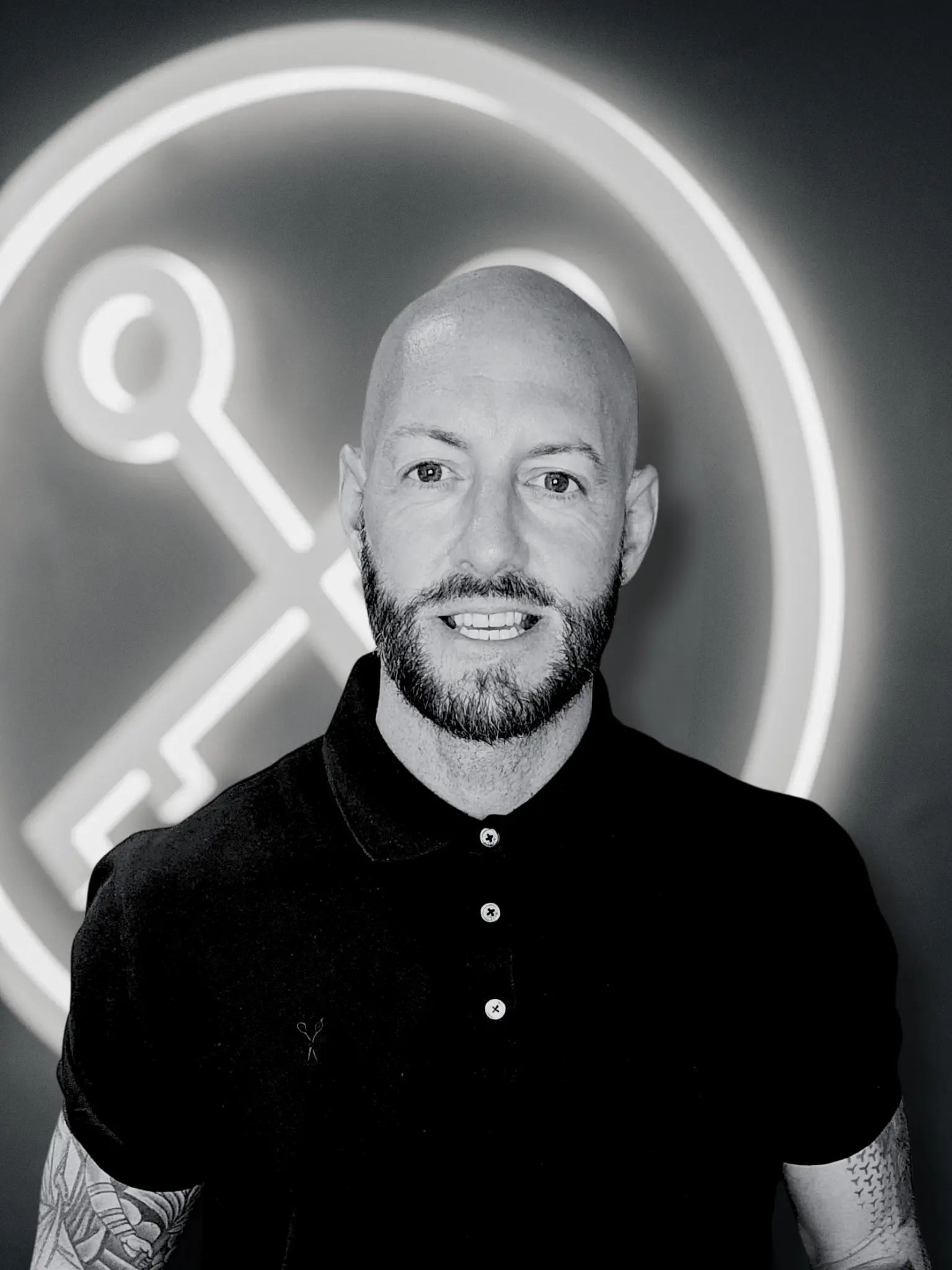 Man with shaved head and beard wearing black polo shirt, standing in front of neon key symbol