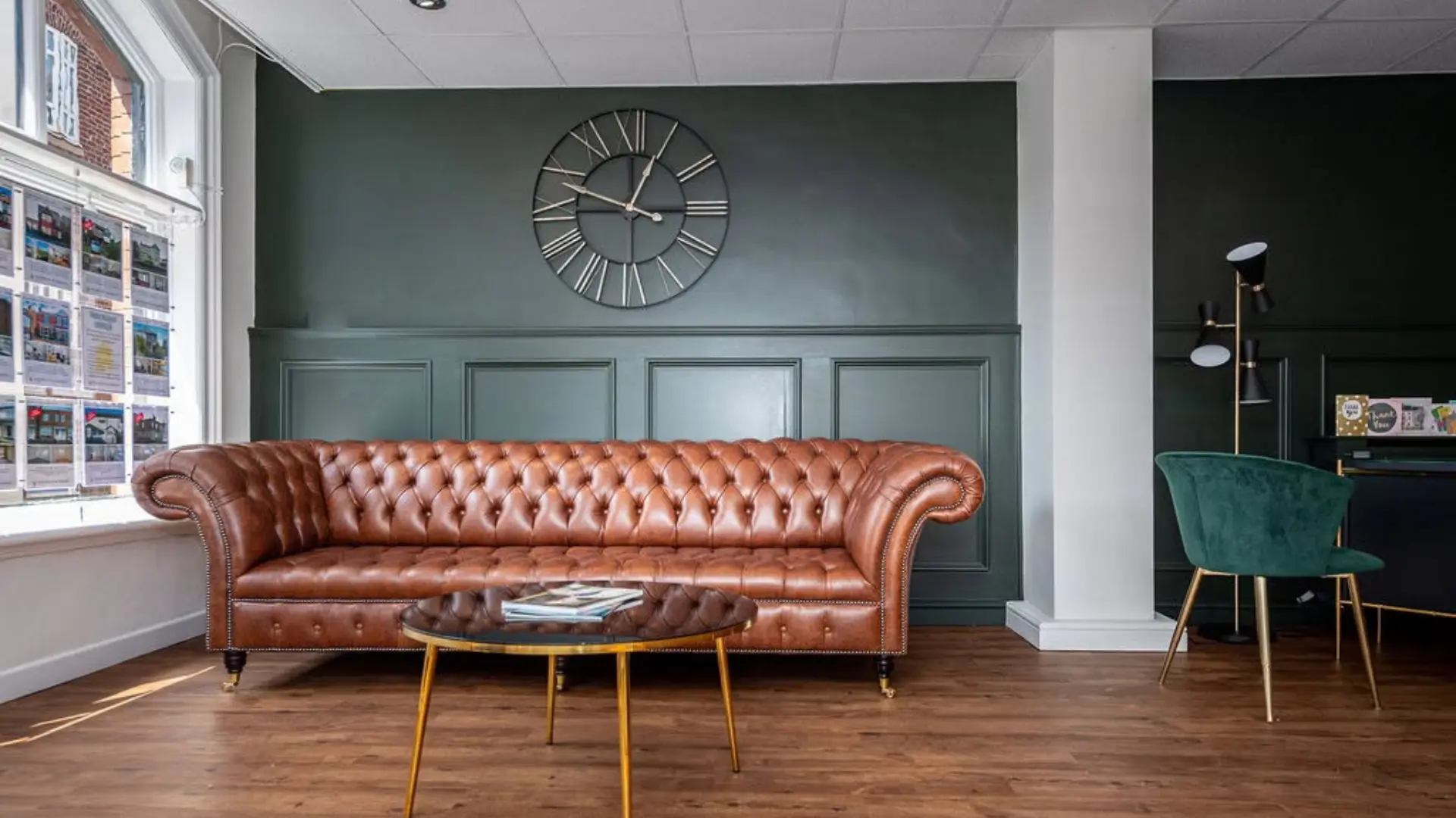 Estate agency office interior with brown leather Chesterfield sofa, gold-legged coffee table, green accent chair, and property listings on window display