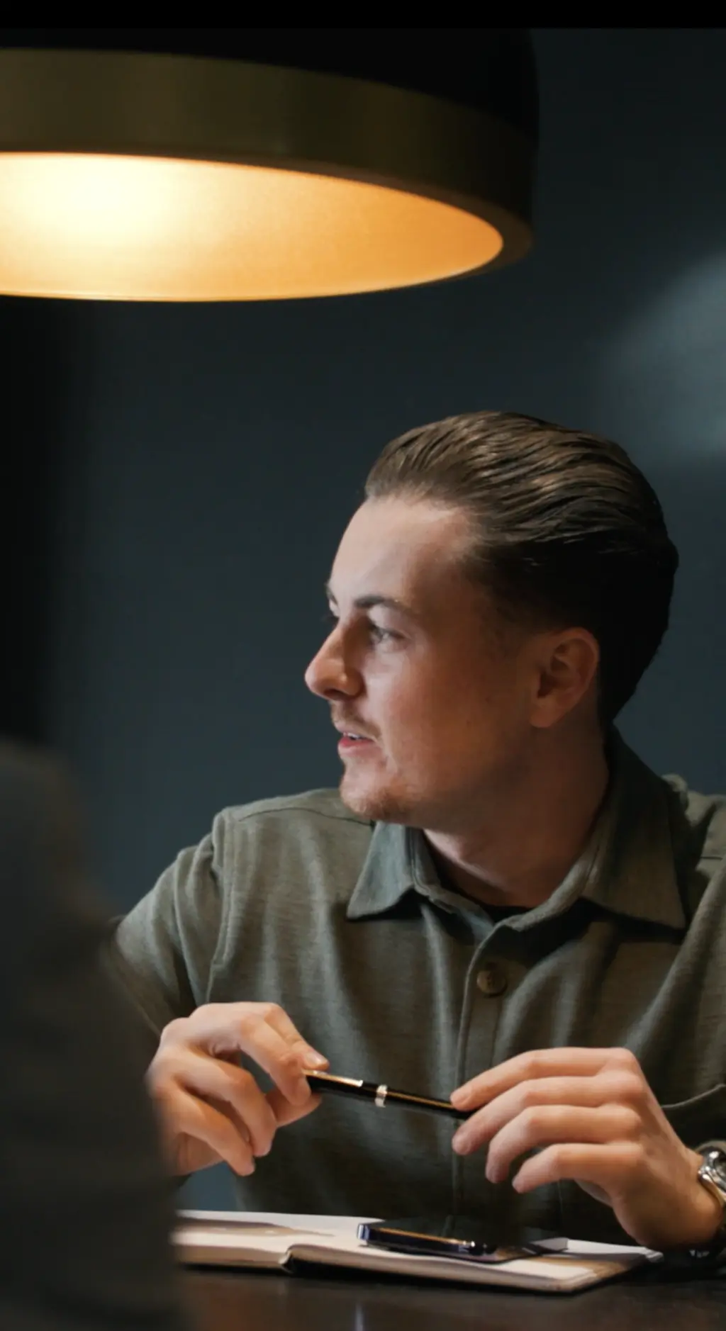 Man in olive shirt holding a pen, seated at a desk under a warm ceiling light