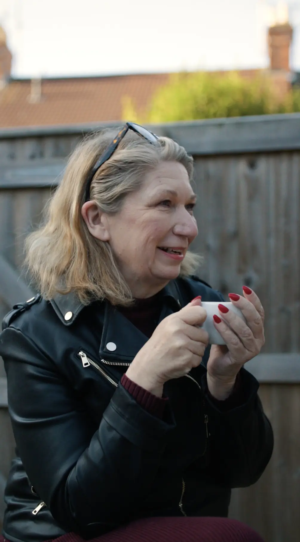 Woman in black leather jacket holding a white mug, standing outdoors with a brick building and fence in the background