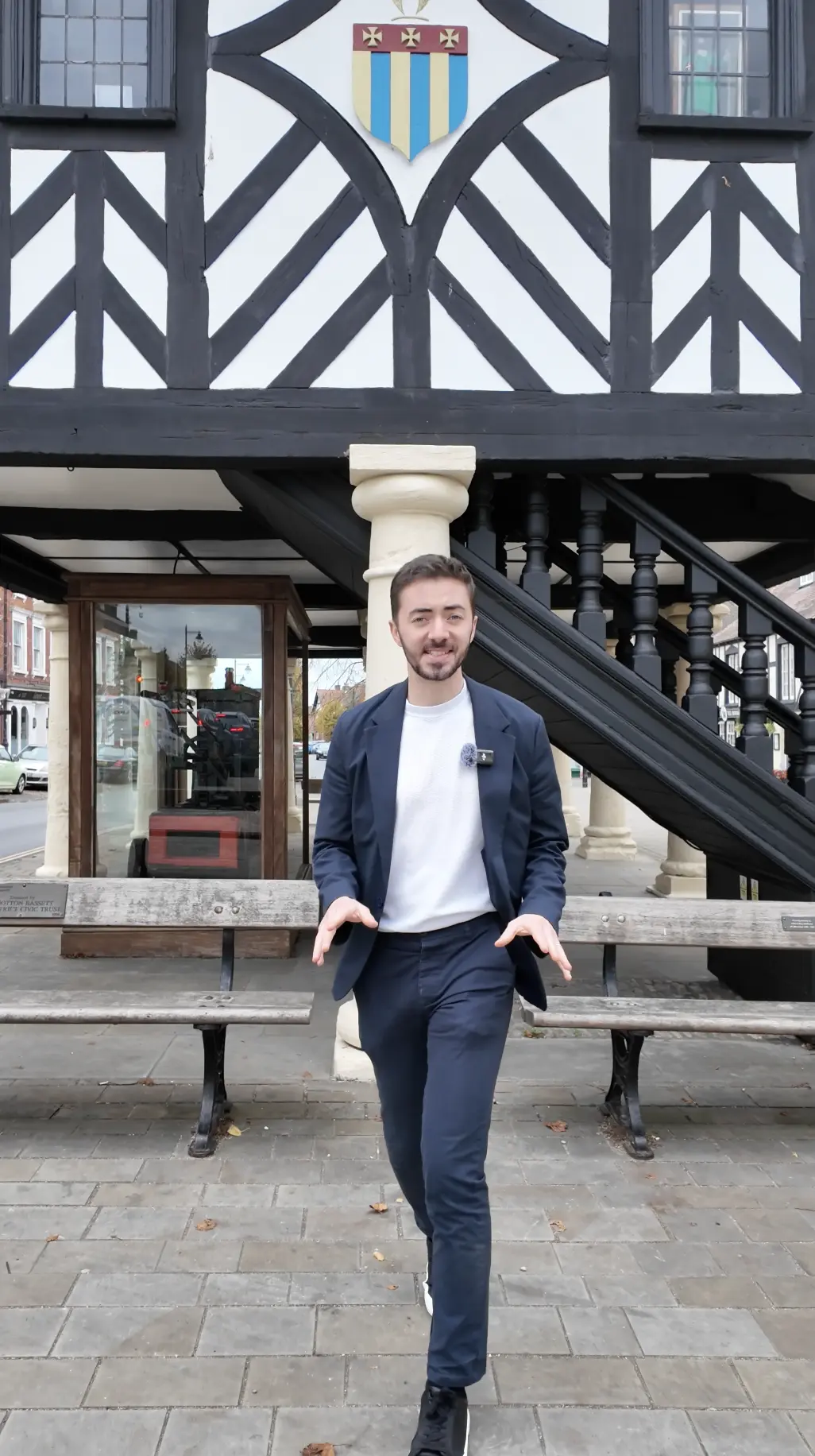 Man in navy blazer and white shirt standing outside a half-timbered building with a heraldic shield above