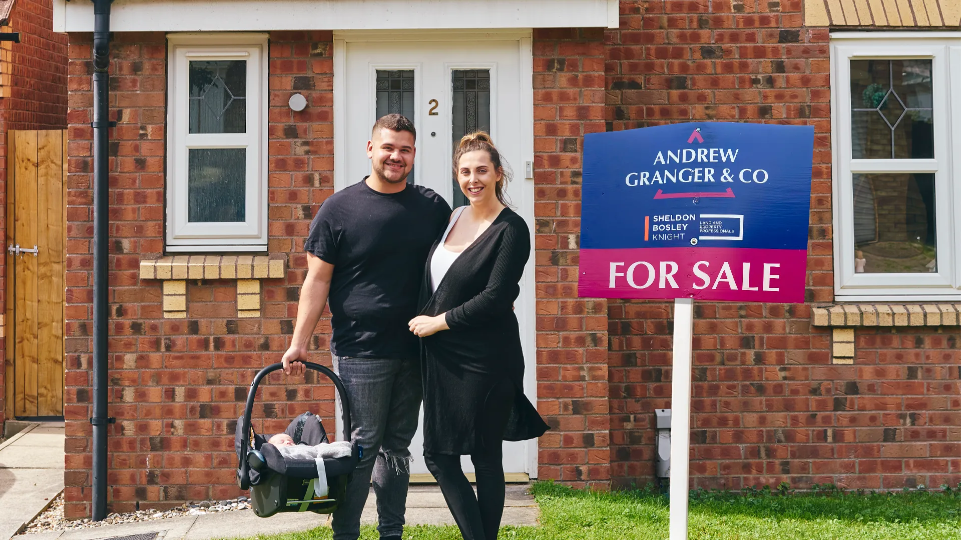 A couple stand on their new front lawn next to an Andrew Granger sold board