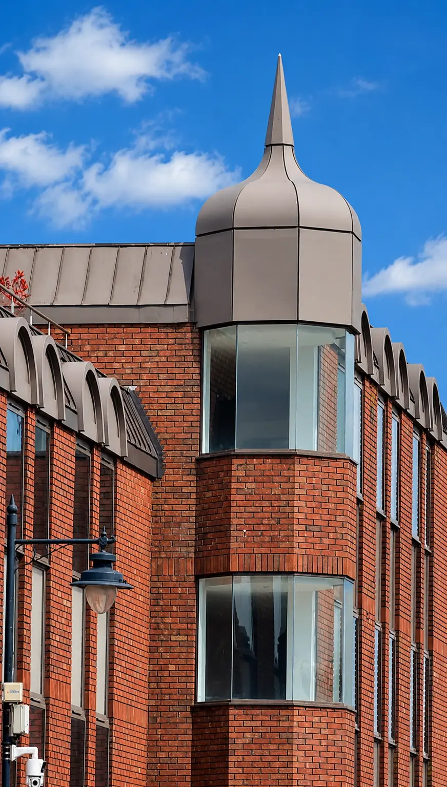 Red brick building with distinctive domed tower featuring a pointed spire, curved bay windows, and mansard roof against blue sky