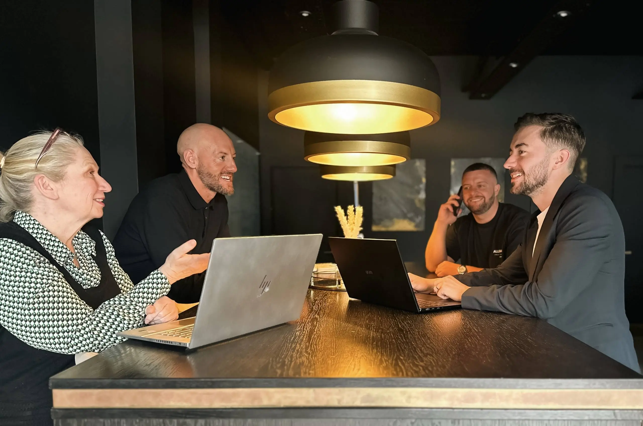 Four people seated at a wooden table with laptops, engaged in conversation in a modern office space with tiered gold pendant lighting