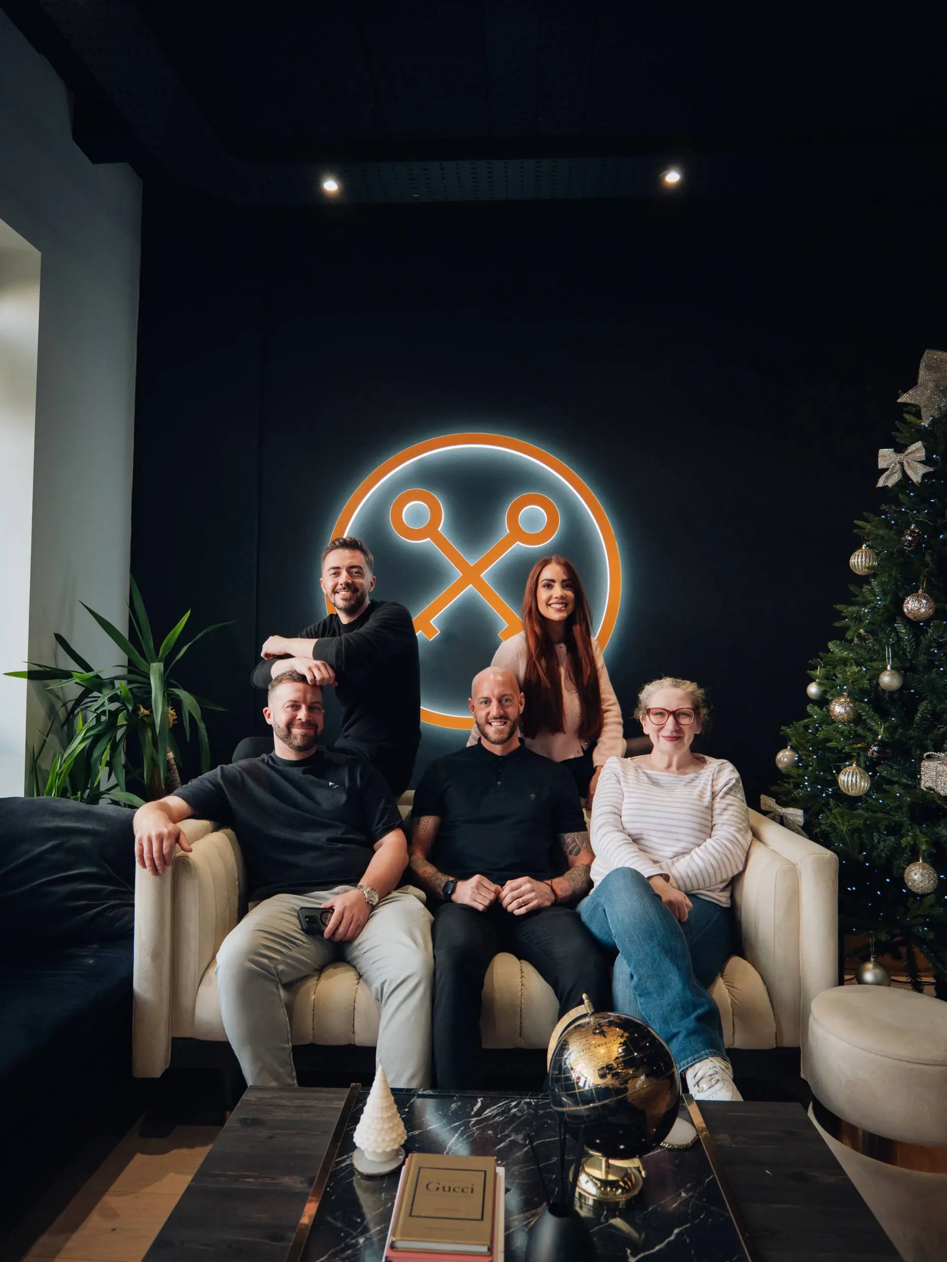 Five people posing on a cream sofa in front of an orange neon X logo, with a decorated Christmas tree to the right and potted plant to the left