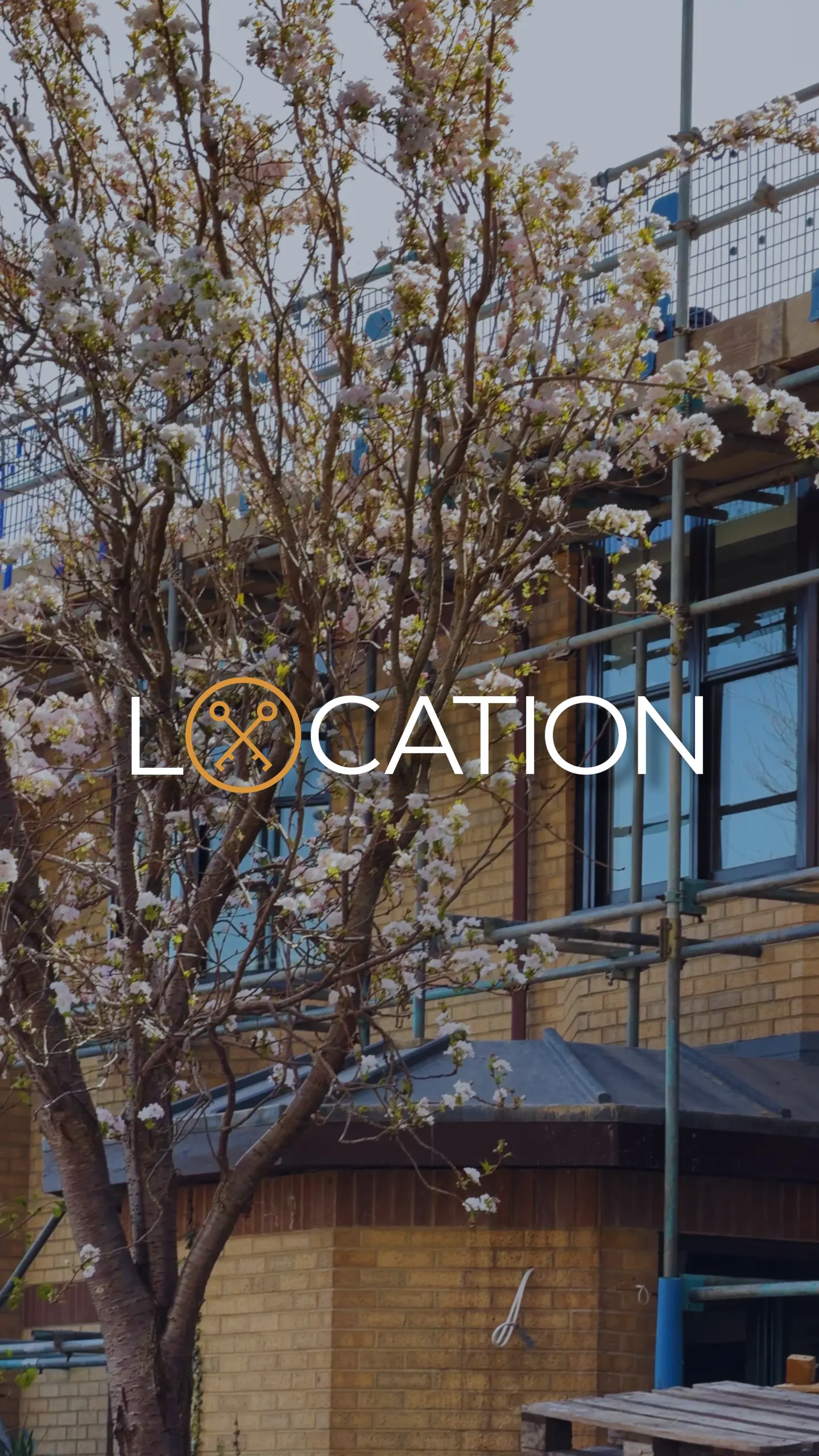 Flowering magnolia tree in front of a brick building with blue-framed windows and metal railings, with text overlay reading 'LOCATION' and a compass logo