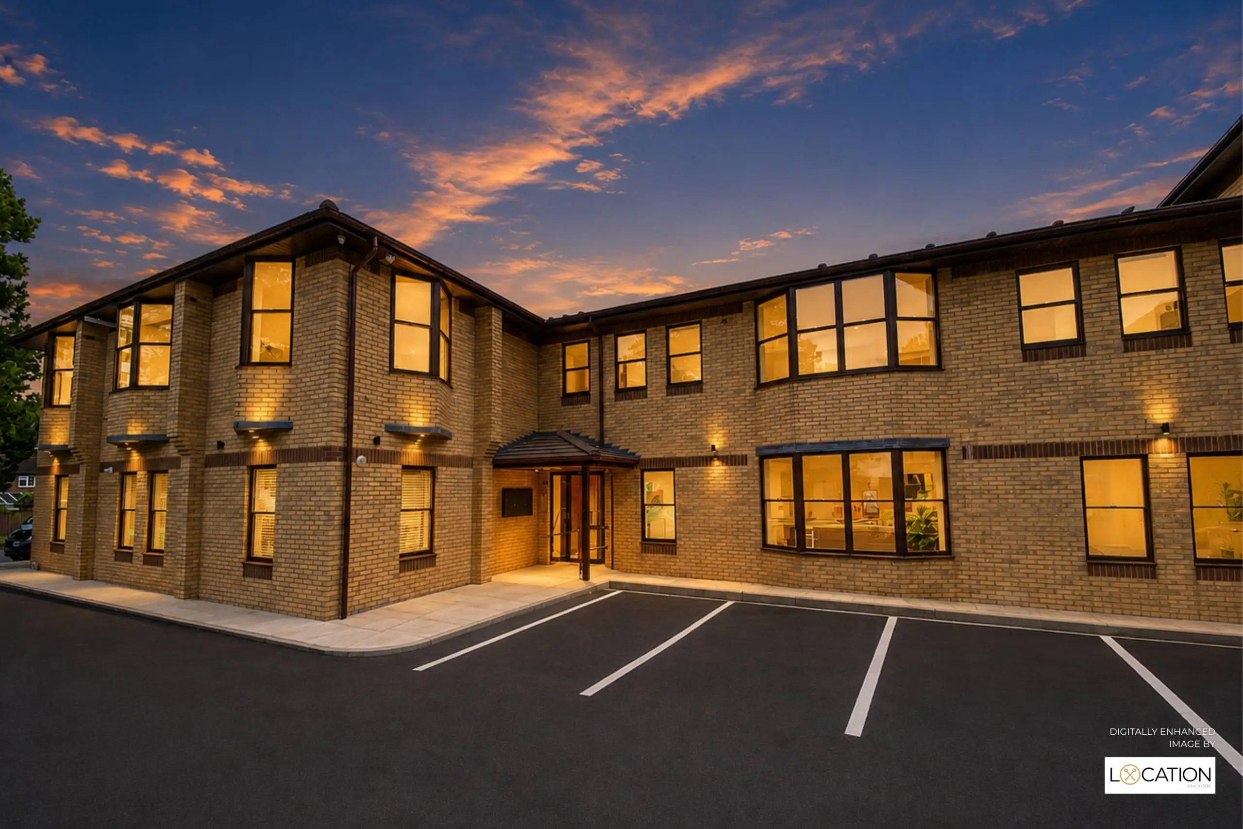 Modern brick building with lit windows at dusk, featuring a central entrance, parking spaces in foreground, and dramatic orange sky