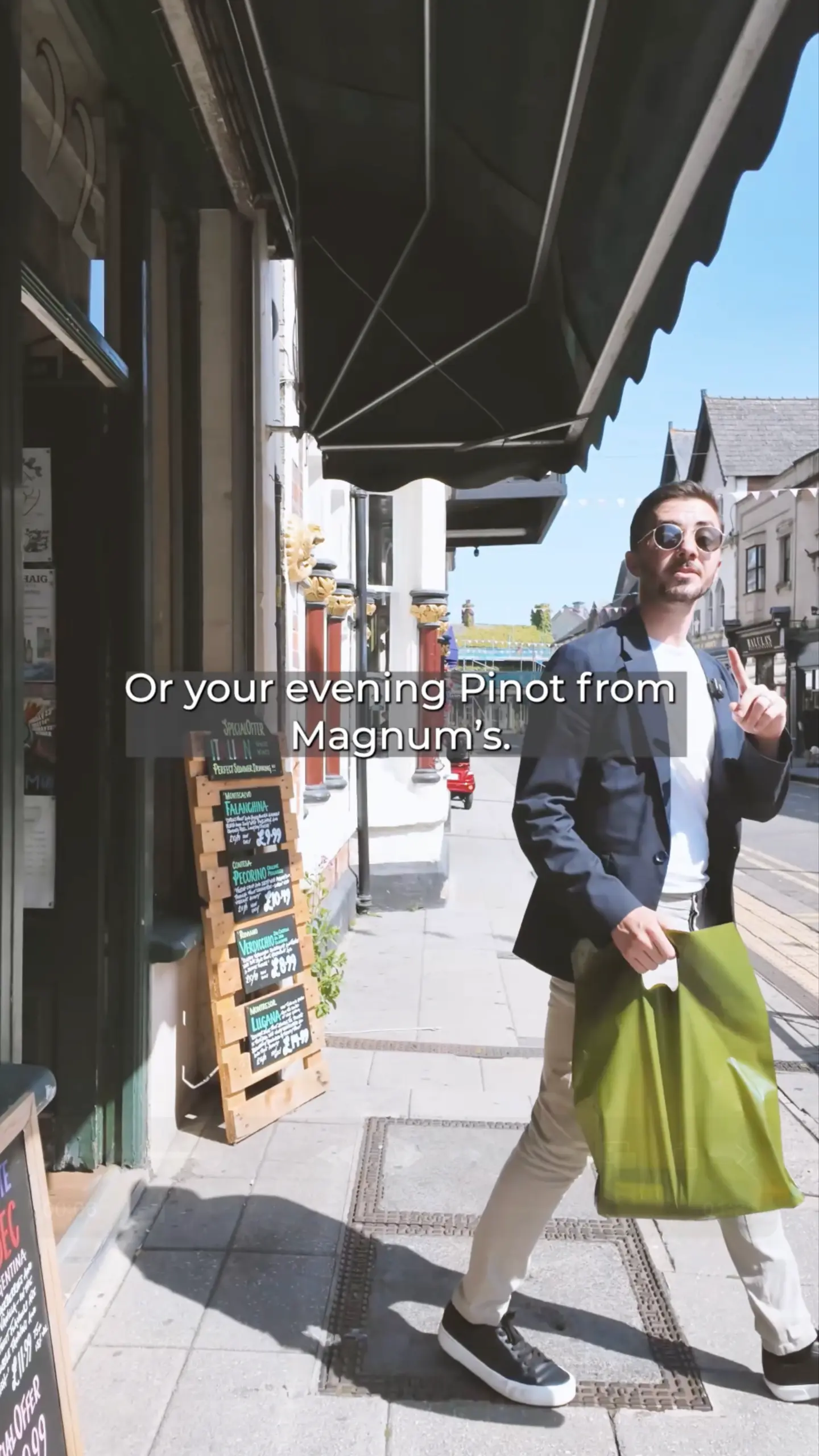 Person wearing sunglasses and navy blazer standing on a street under a retractable awning, with text overlay reading 'Or your evening Pinot from Magnum's.'