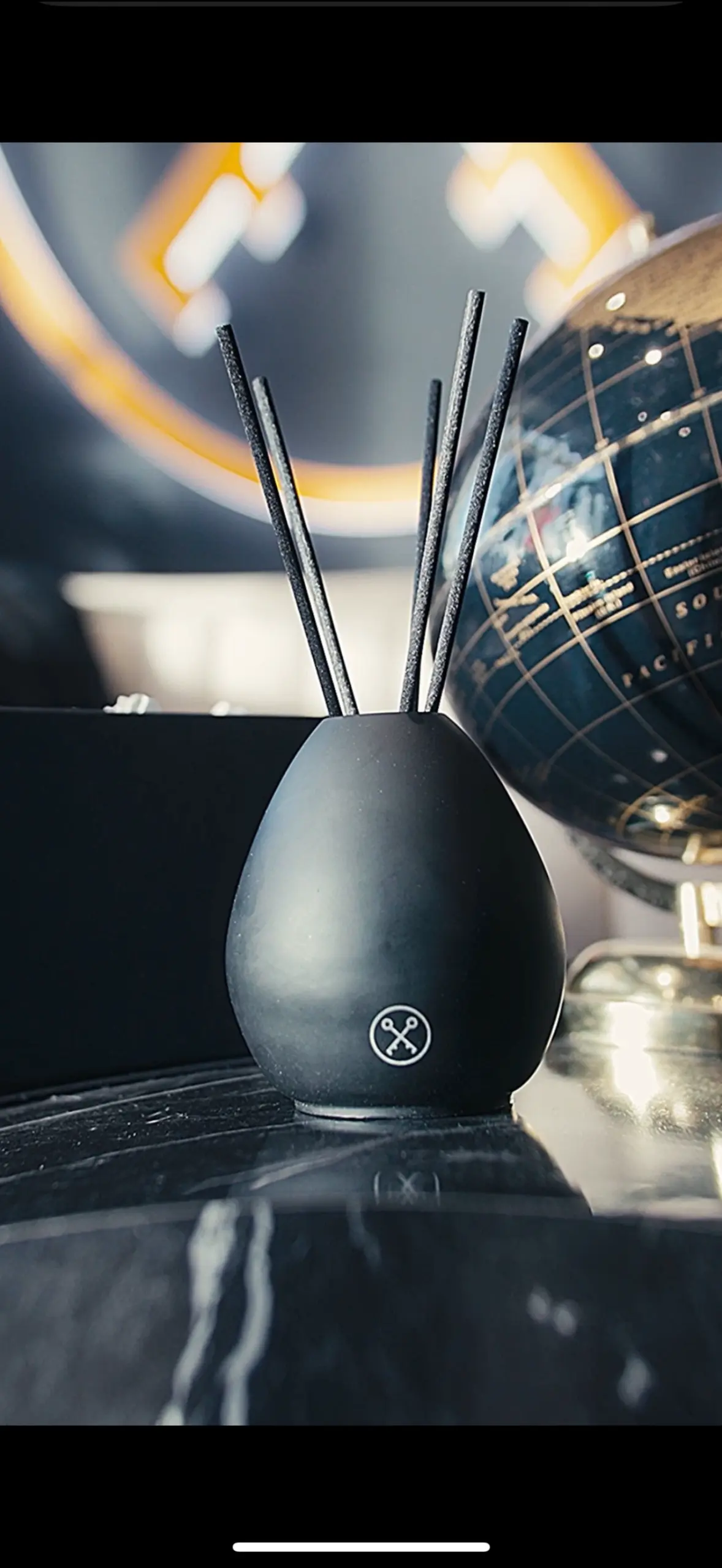 Black egg-shaped diffuser with four black sticks on a dark surface, beside a decorative globe, with warm blurred lighting in the background