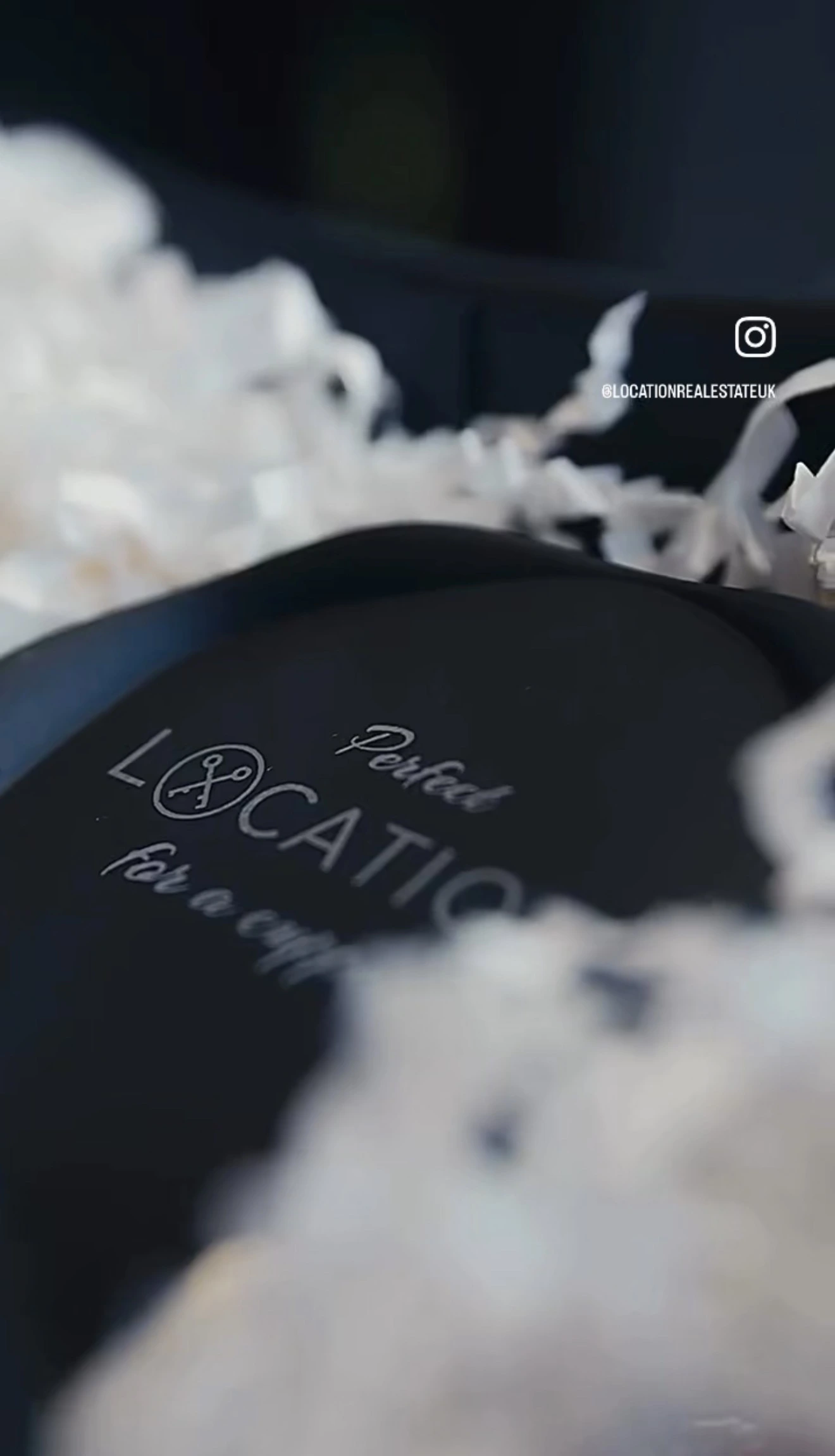 Close-up of a black plate with engraved text reading 'LOCATION' and 'Petrol' with white flowers blurred in the background
