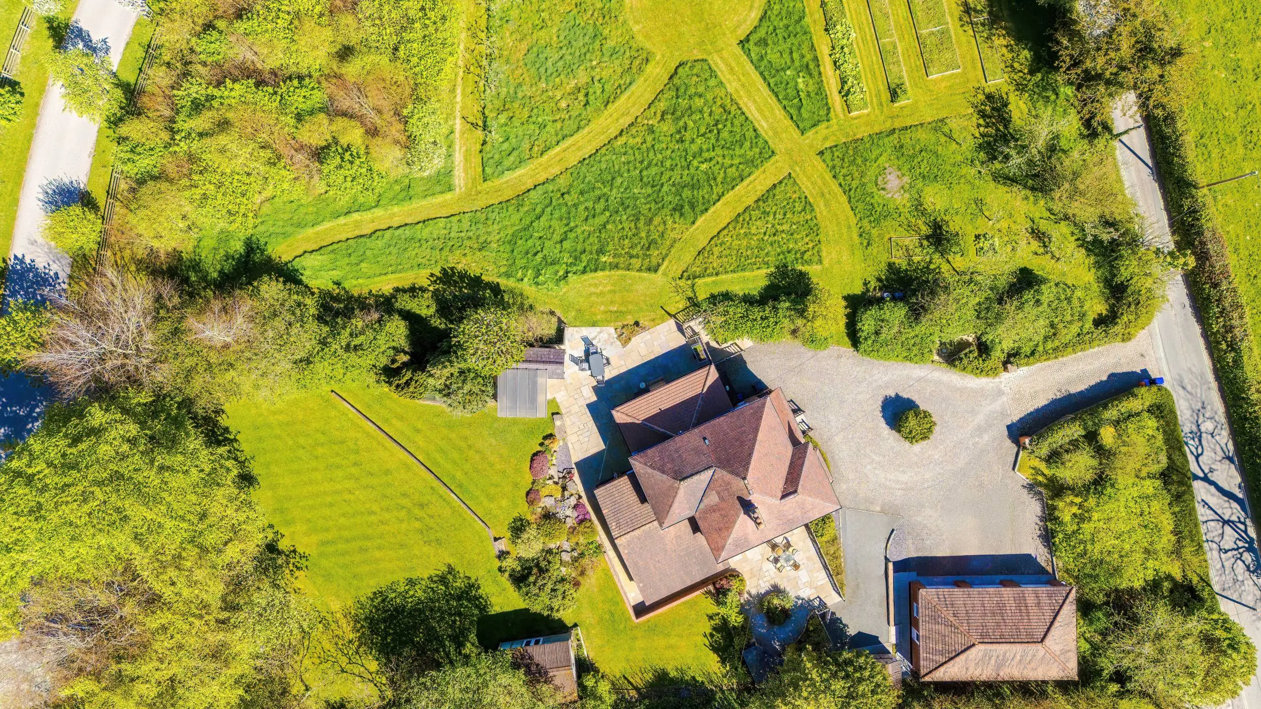 Aerial view of a residential property with terracotta roof tiles surrounded by mature trees and green lawn, with a driveway and outbuildings visible